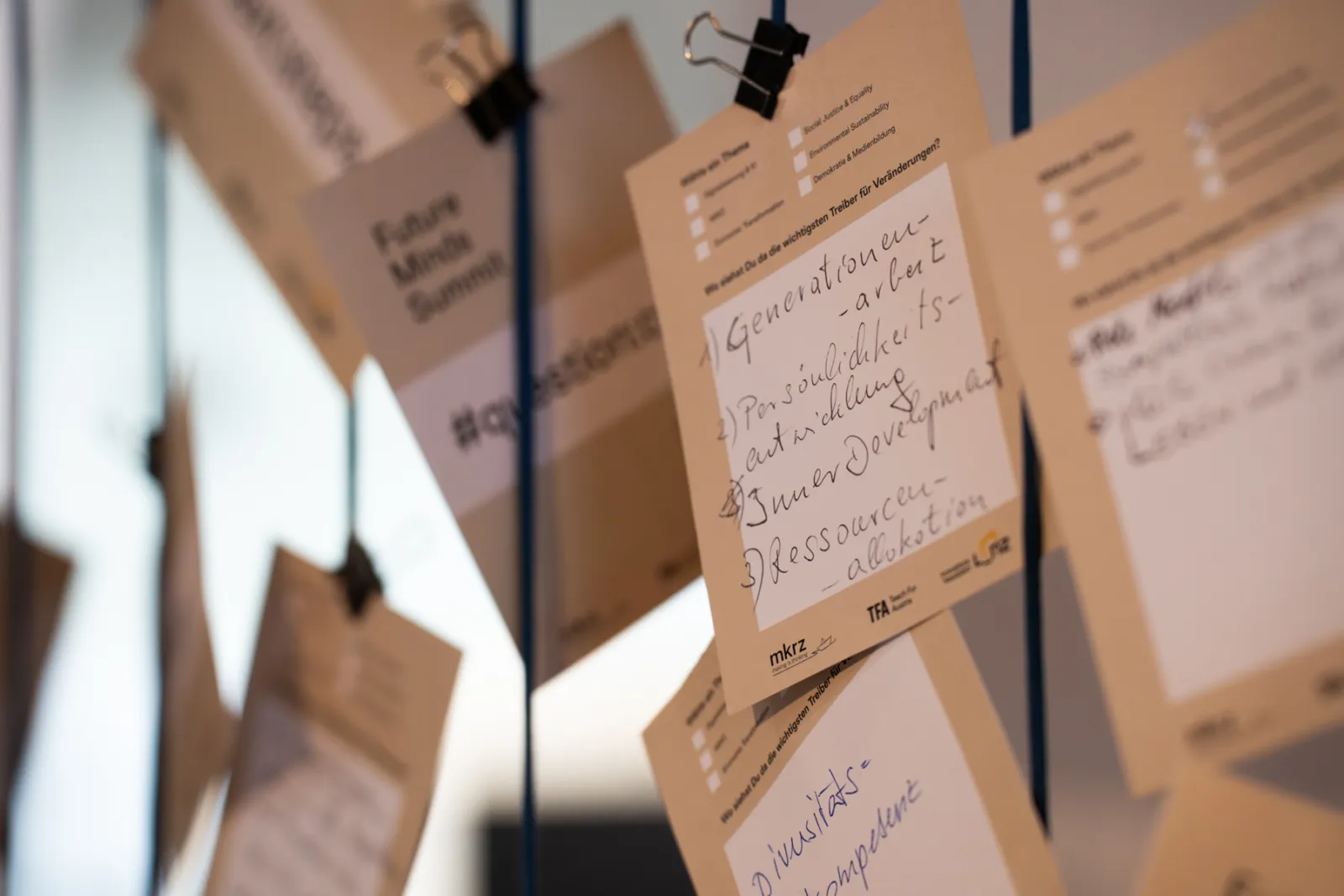 Mehrere beigefarbene Notizzettel hängen an Clips, befestigt an dünnen Schnüren; unscharfer Innenraum im Hintergrund.