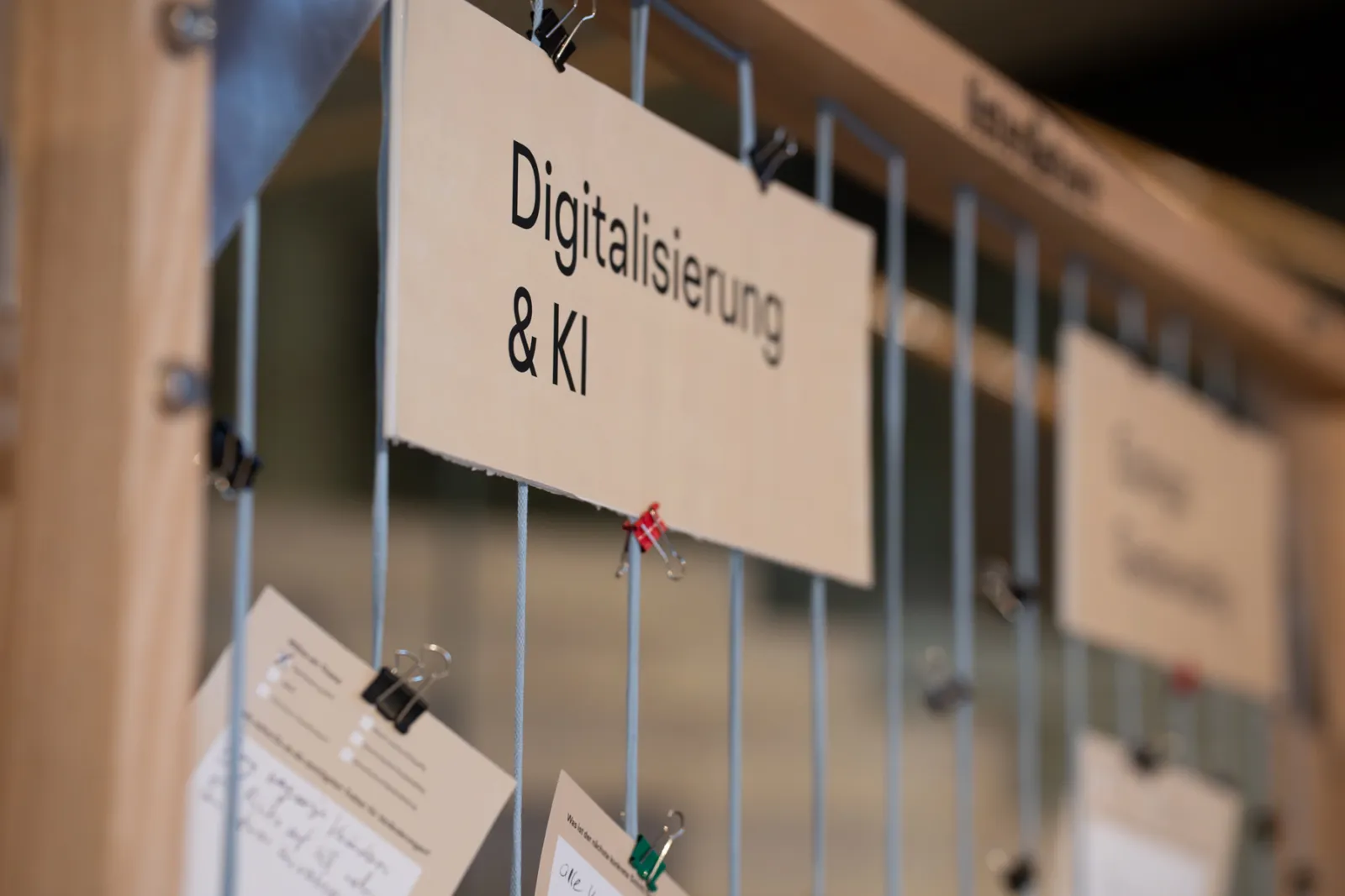 Schild mit der Aufschrift Digitalisierung & KI hängt an einem Metallgitter, umgeben von Notizzetteln.