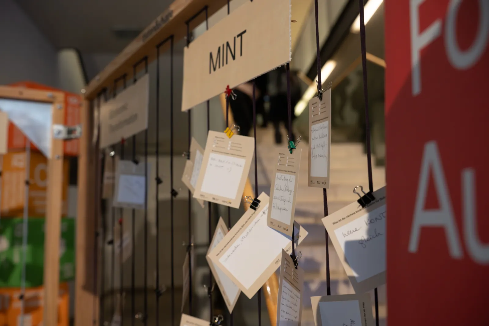 Viele handschriftliche Notizkarten hängen an Schnüren unter dem Schild 'MINT' in einem Ausstellungsbereich.