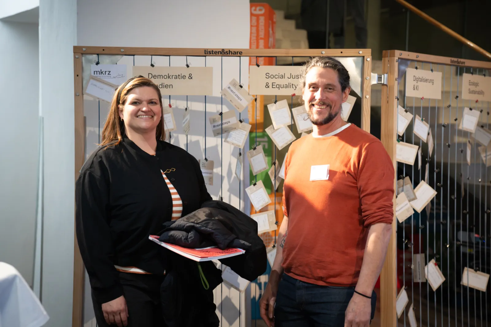 Zwei Personen vor Infowand mit Post-it-Notizen; eine hält Mantel und Unterlagen, andere lächelt; Veranstaltungsumgebung.
