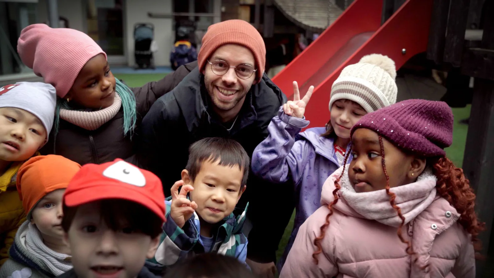 Gruppe von Kindern verschiedener Herkunft um einen Mann mit Brille und orangefarbener Mütze auf Spielplatz; ein Kind macht Peace-Zeichen vor roter Rutsche.