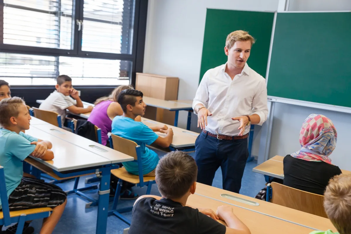 Lehrkraft erklärt Gruppe von Schülern im Klassenzimmer; Tische, Stühle und grüne Tafel im Hintergrund.
