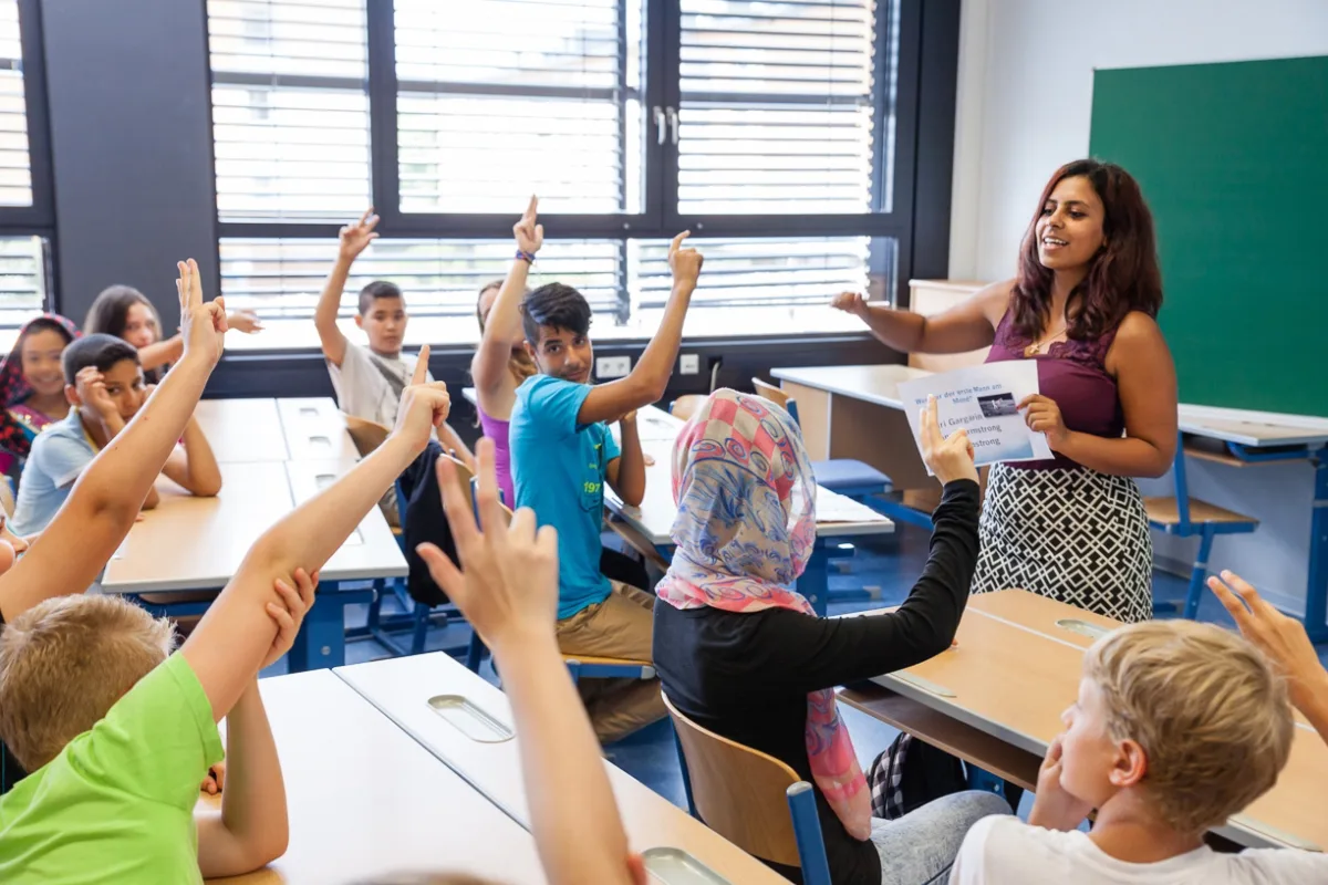 Lehrkraft steht vorne im Klassenraum, erklärt Lernstoff; Lernende heben die Hände, sitzen an Tischen neben großen Fenstern.