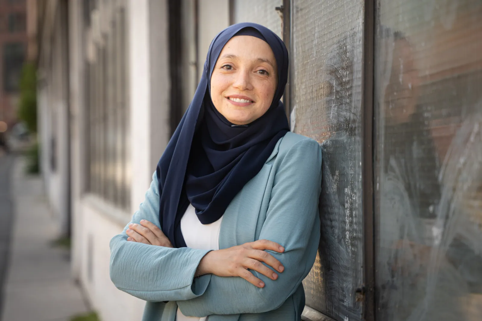 Frau im dunkelblauen Hijab, hellblauen Blazer, lehnt an Glasfassade am Gehsteig, Arme verschränkt, lächelt.