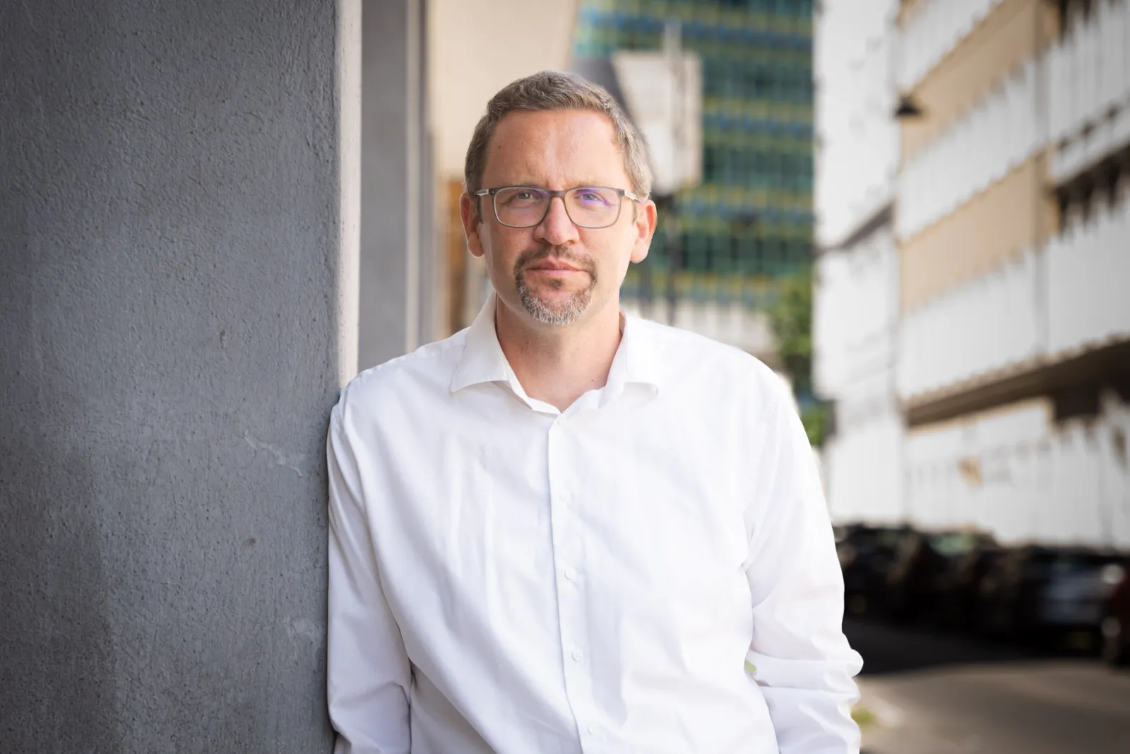 Mann mit Brille in weißem Hemd lehnt an grauer Wand, urbane Straßenszene im Hintergrund.