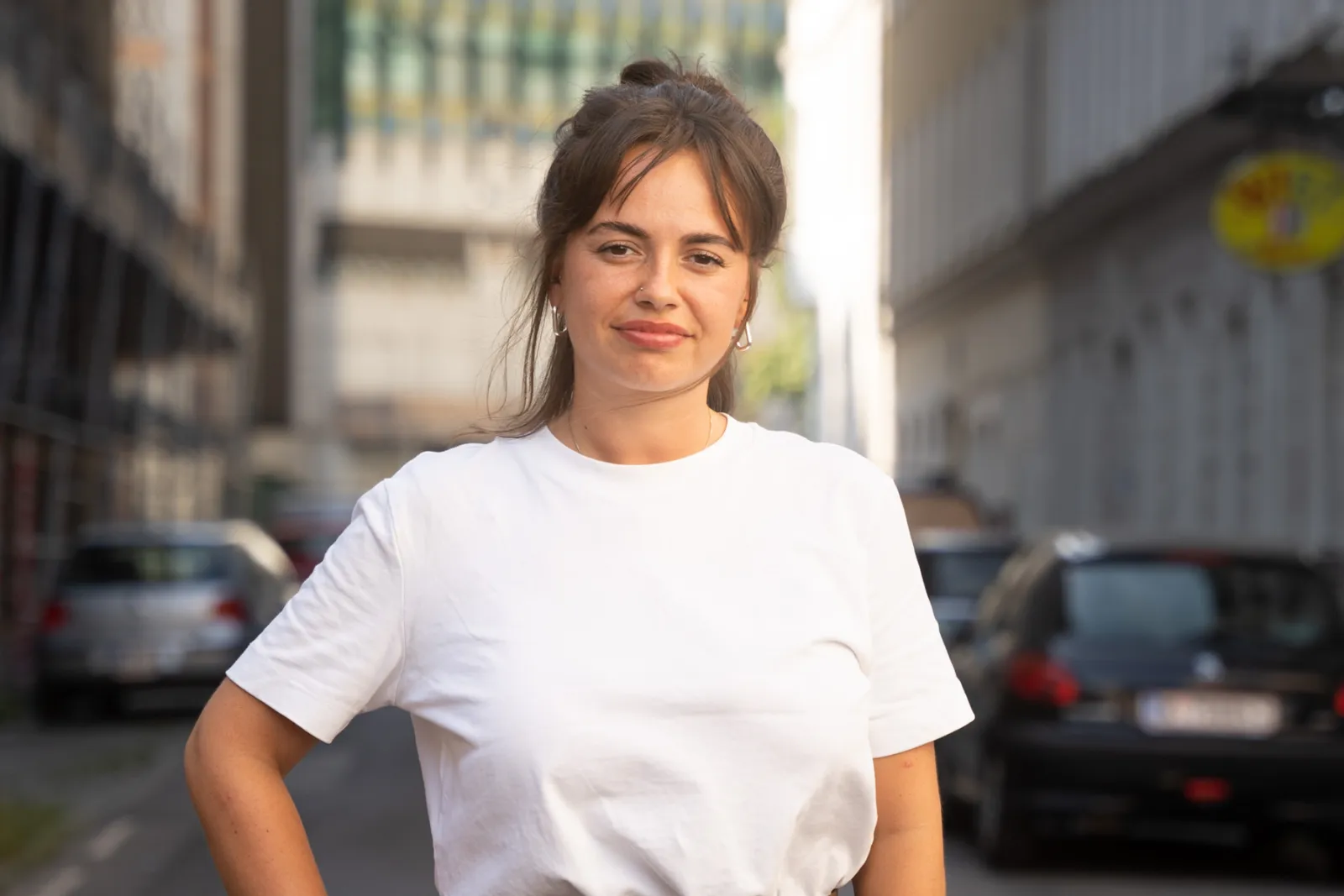 Person in weißem T-Shirt steht mit Hand auf der Hüfte in einer urbanen Straßenszene; im Hintergrund geparkte Autos.