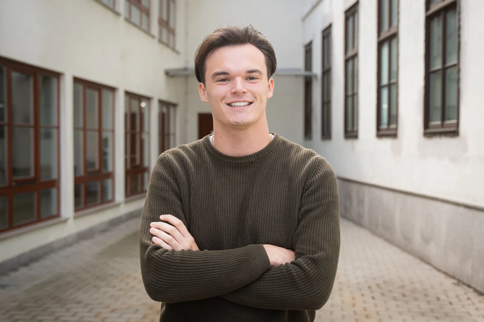 Junge Person lächelt, mit verschränkten Armen, im Innenhof eines Gebäudekomplexes.
