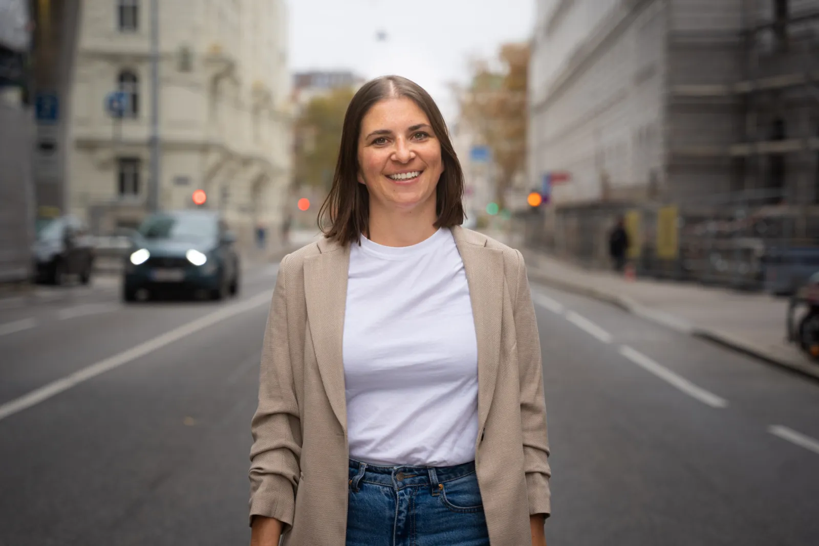 Porträt einer Person in der Mitte einer Stadtstraße, lächelnd; Beige Blazer, weißes T-Shirt, Jeans; urbaner Hintergrund mit Gebäuden und Verkehr.