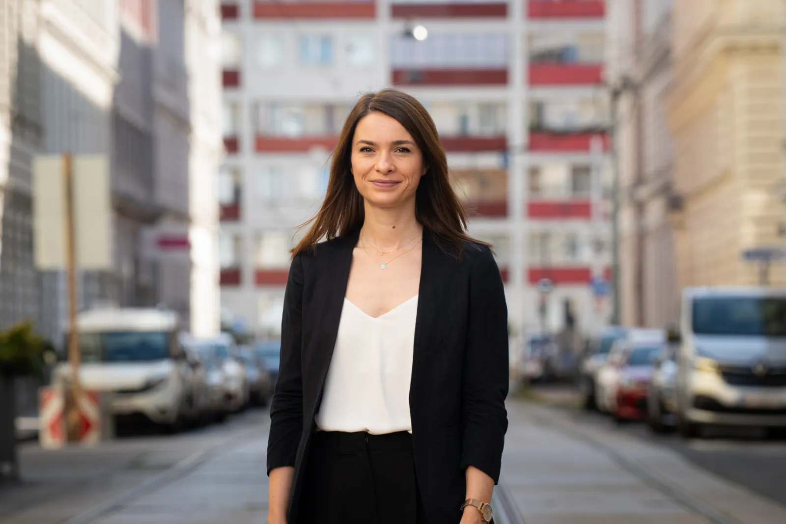 Person in schwarzem Blazer und weißem Top steht lächelnd in einer urbanen Straßenszene mit Autos.