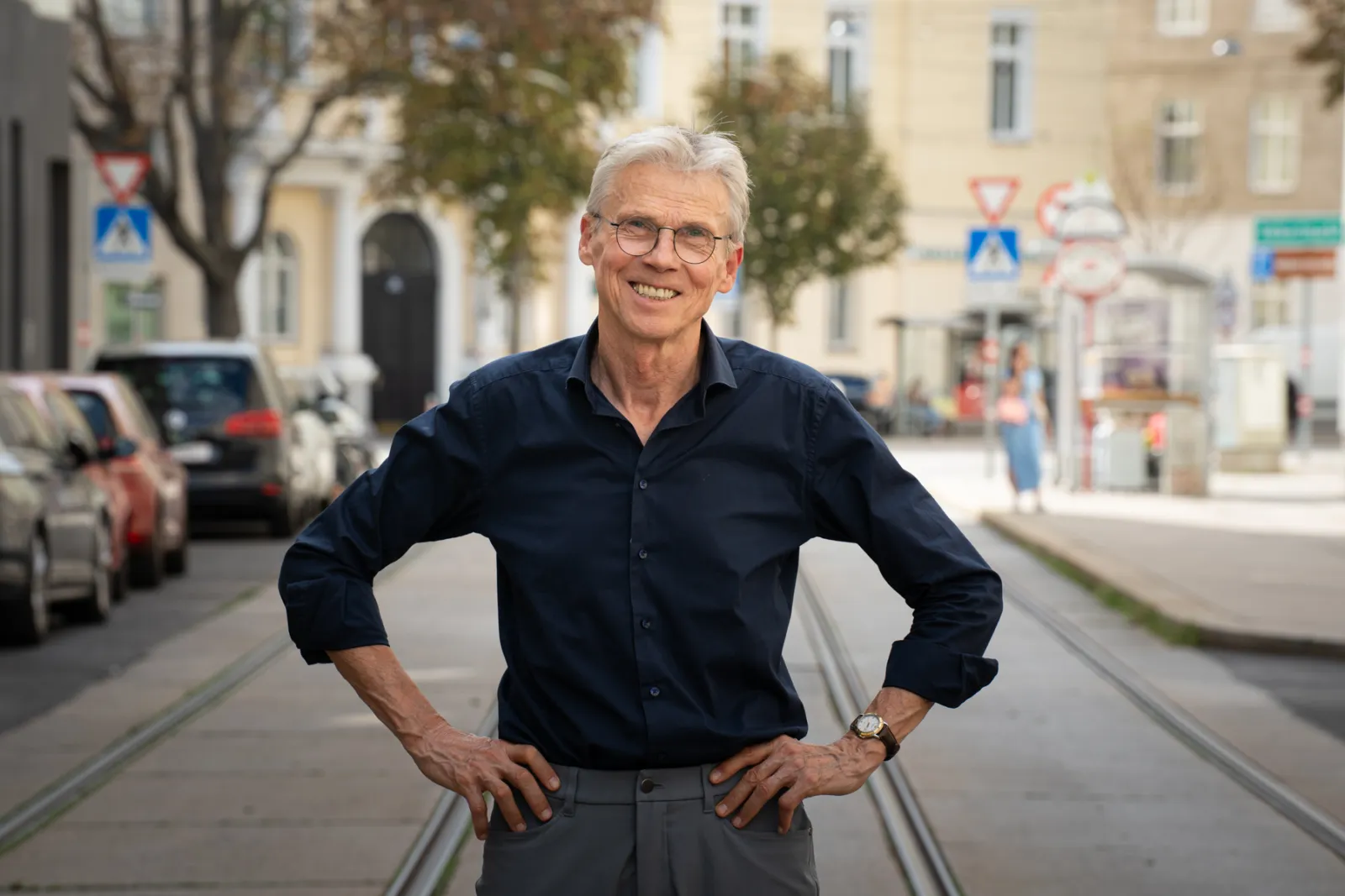 Ältere Person mit Brille steht auf einer Straße mit Tramgleisen, Hände in den Hüften, lächelnd; urbane Umgebung.