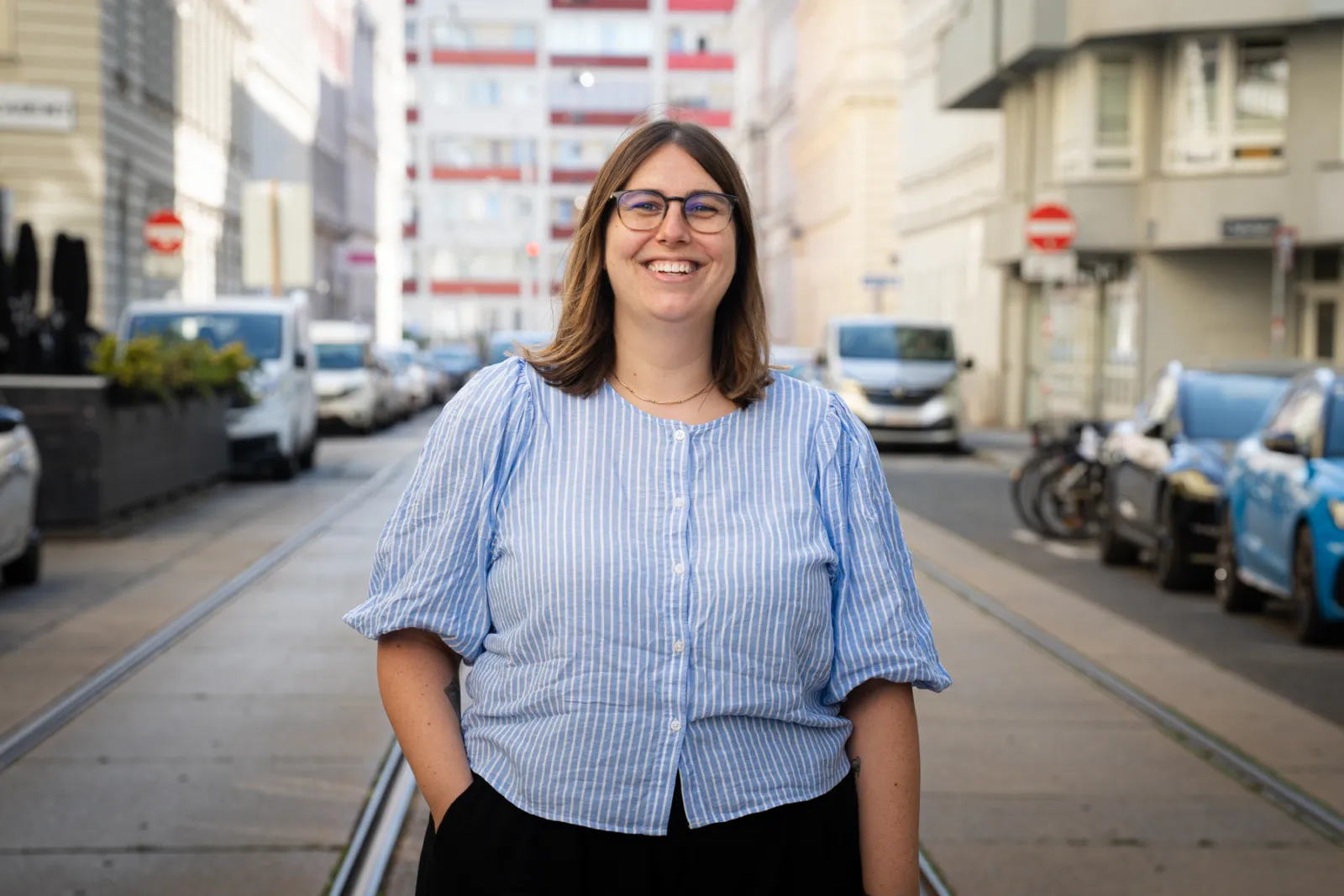 Person mit Brille lächelt, trägt blau-weiß gestreifte Bluse, steht auf einer urbanen Straße mit Schienen.