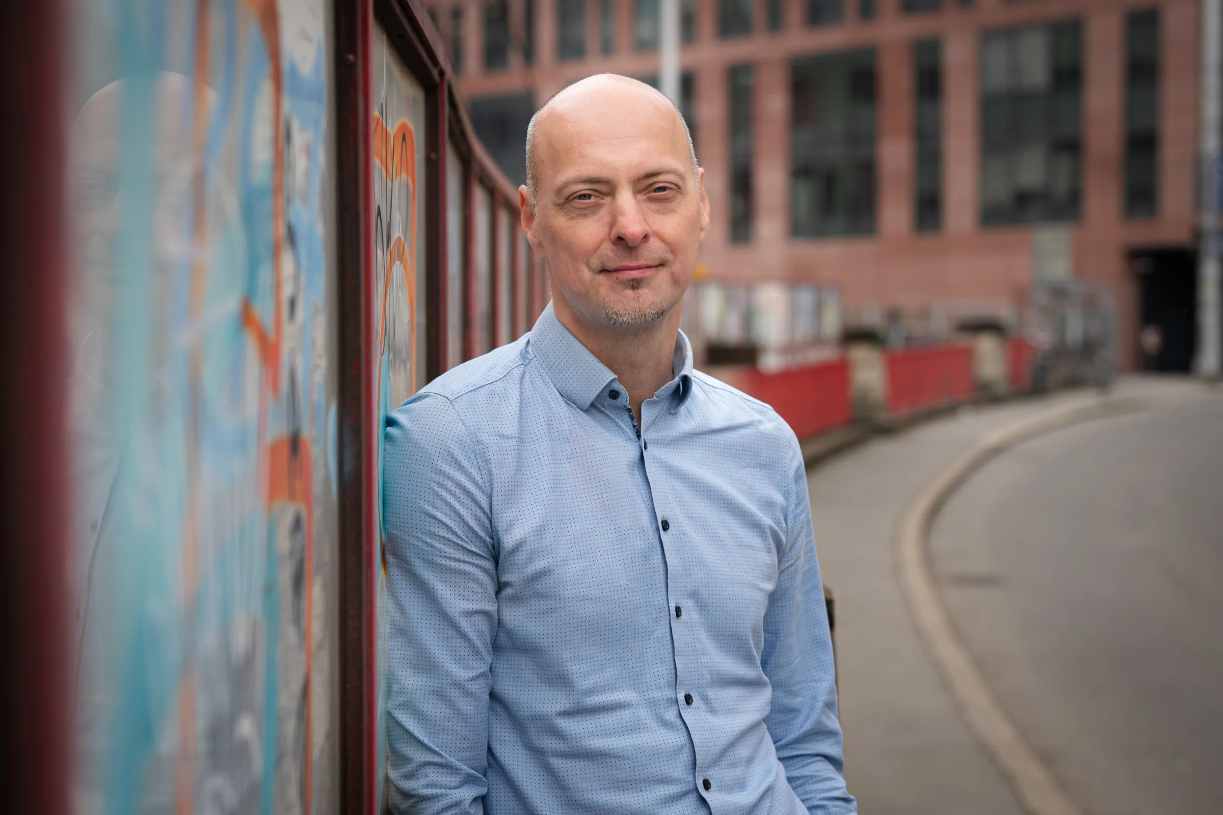 Person lehnt an Graffitimauer in urbaner Straßenszene; trägt hellblaues Hemd, Backsteingebäude im Hintergrund.