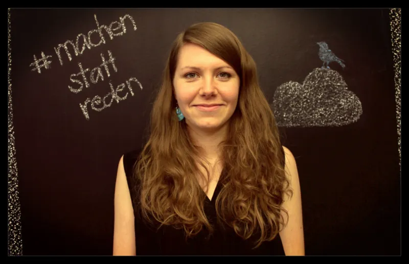 Porträt einer jungen Frau mit langen lockigen braunen Haaren, schwarzes Oberteil, vor Kreidetafel mit herzförmiger Wolke und Vogel.