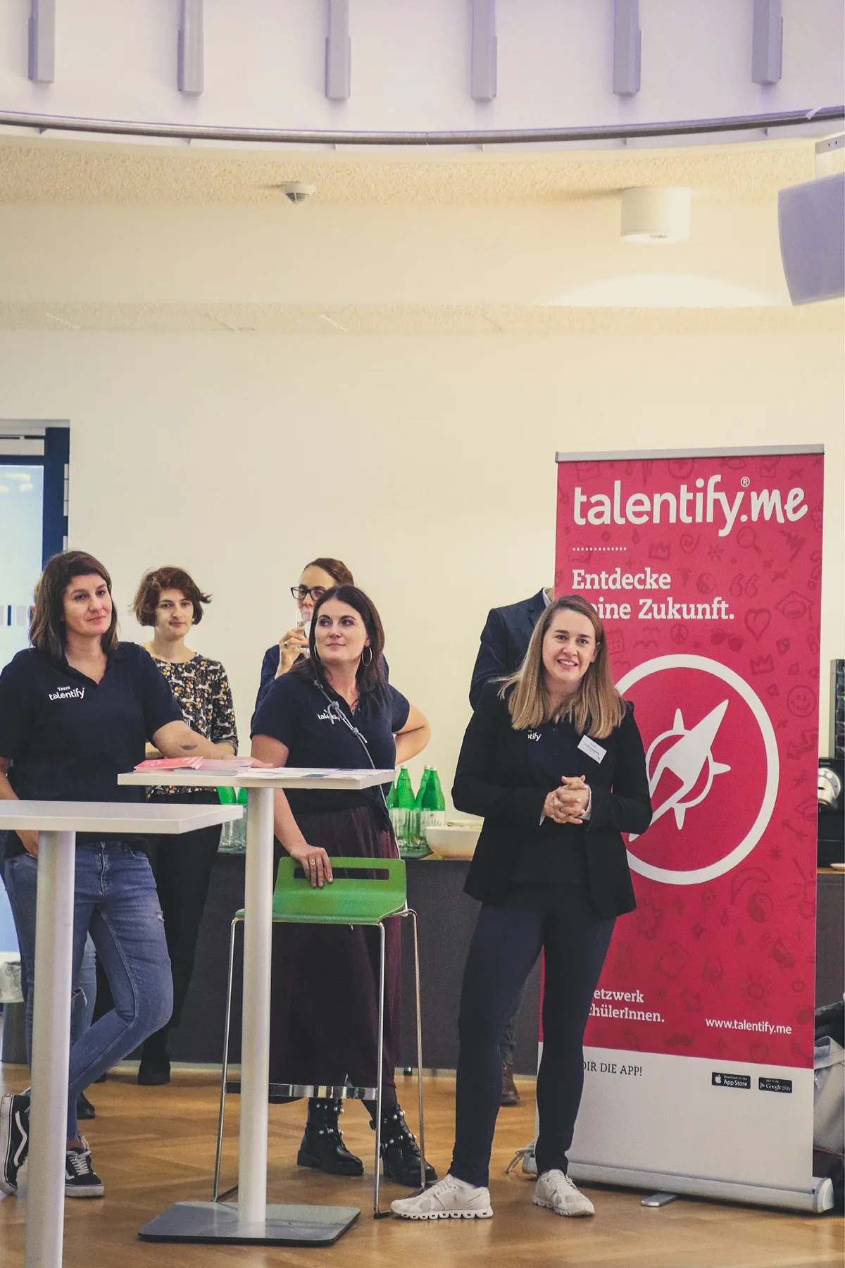 Mehrere Frauen stehen hinter einem Stehtisch neben einem pinken Banner mit 'talentify.me' in einem Veranstaltungsraum.