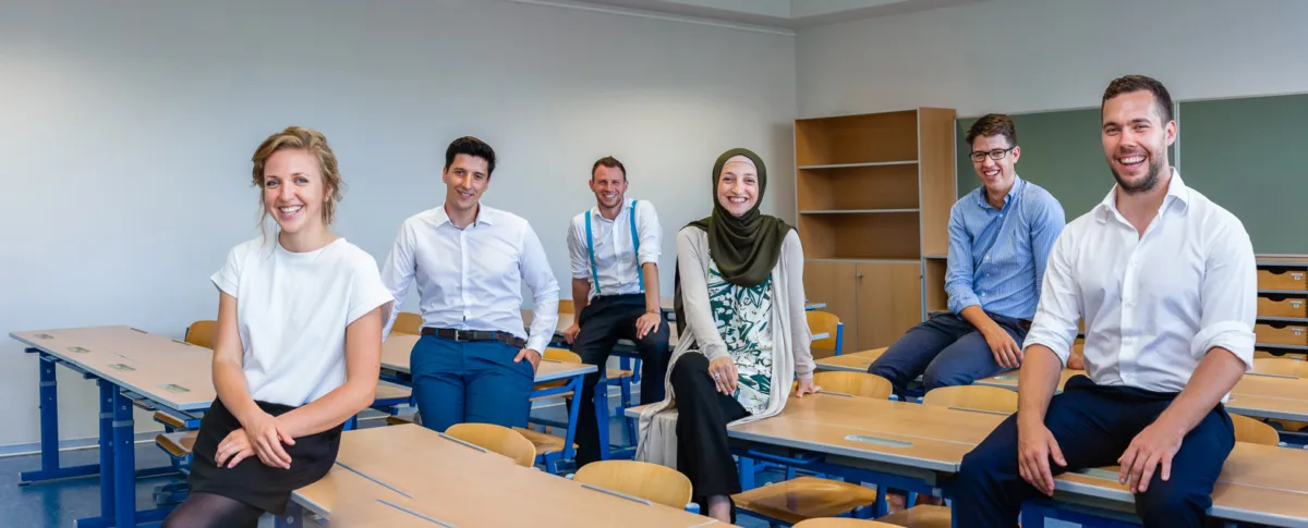 Sechs Personen sitzen lächelnd auf Schreibtischen in einem Klassenraum.