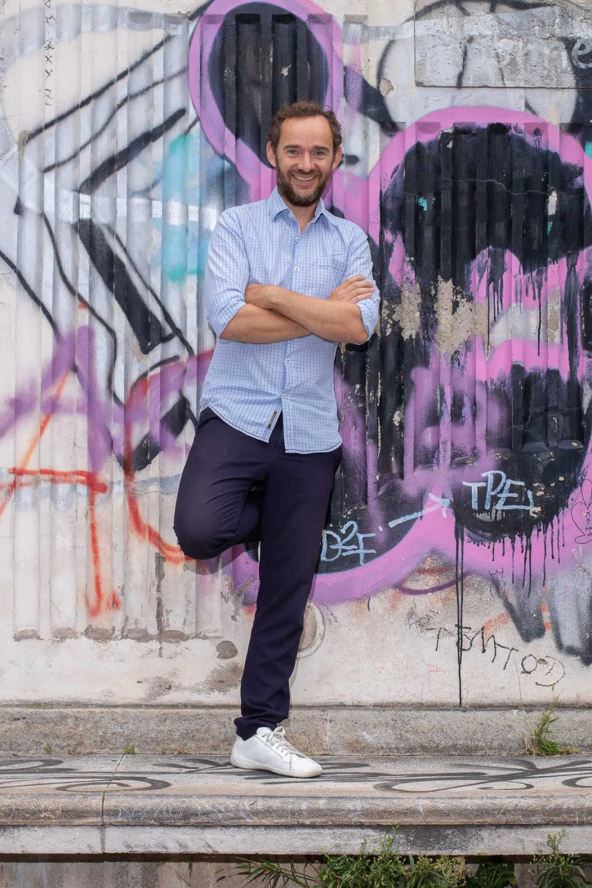 Person steht vor Graffiti-Wand, Arme verschränkt, Bein auf einer Stufe abgestützt; hellblaues kariertes Hemd, dunkle Hose, weiße Sneaker.