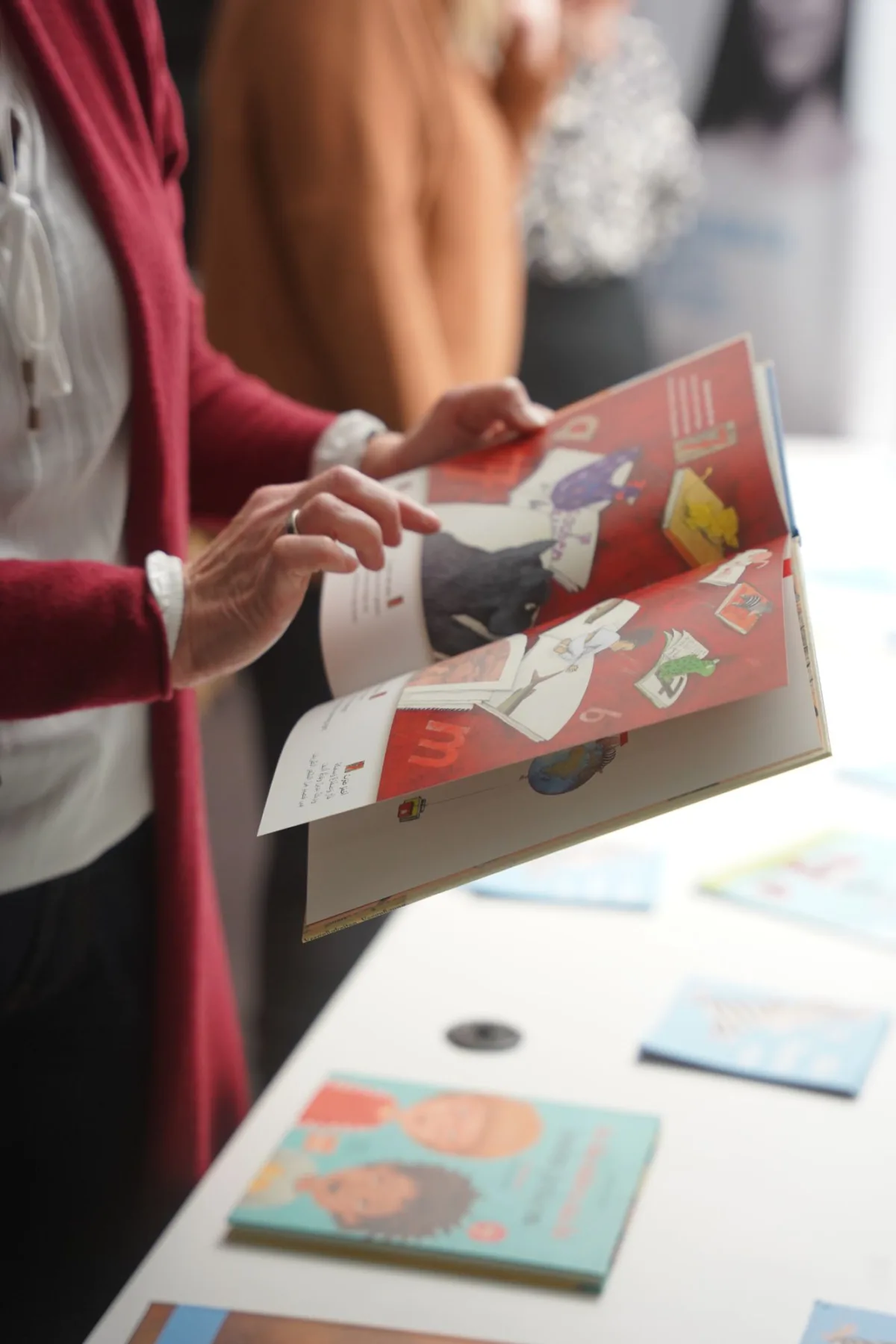 Person blättert durch ein buntes Magazin auf einem Ausstellungstisch; im Hintergrund weitere Besucher.