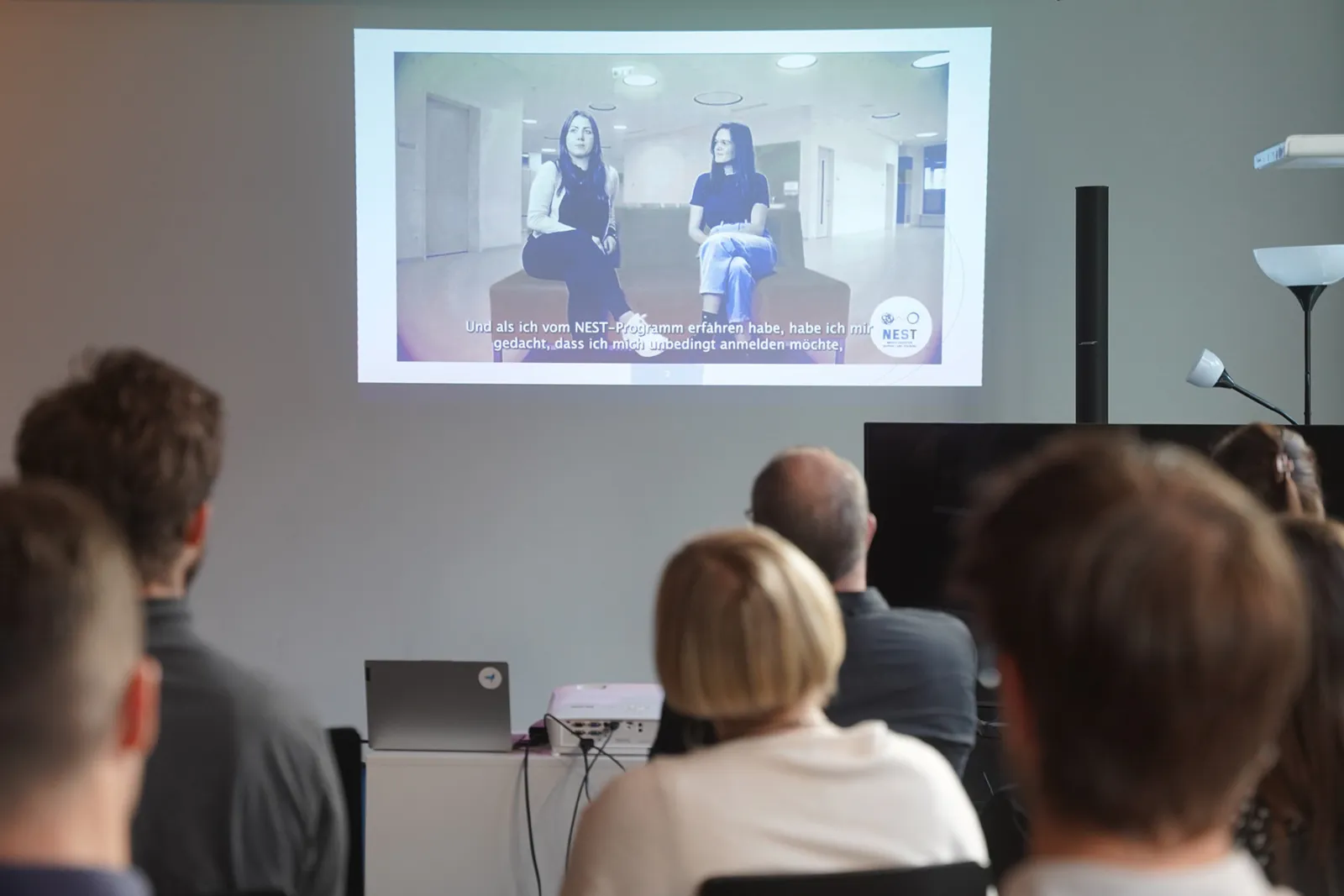 Projektion zeigt zwei Frauen, die in einem hellen Seminarraum sitzen; zuschauendes Publikum, Laptop und Beamer im Vordergrund.