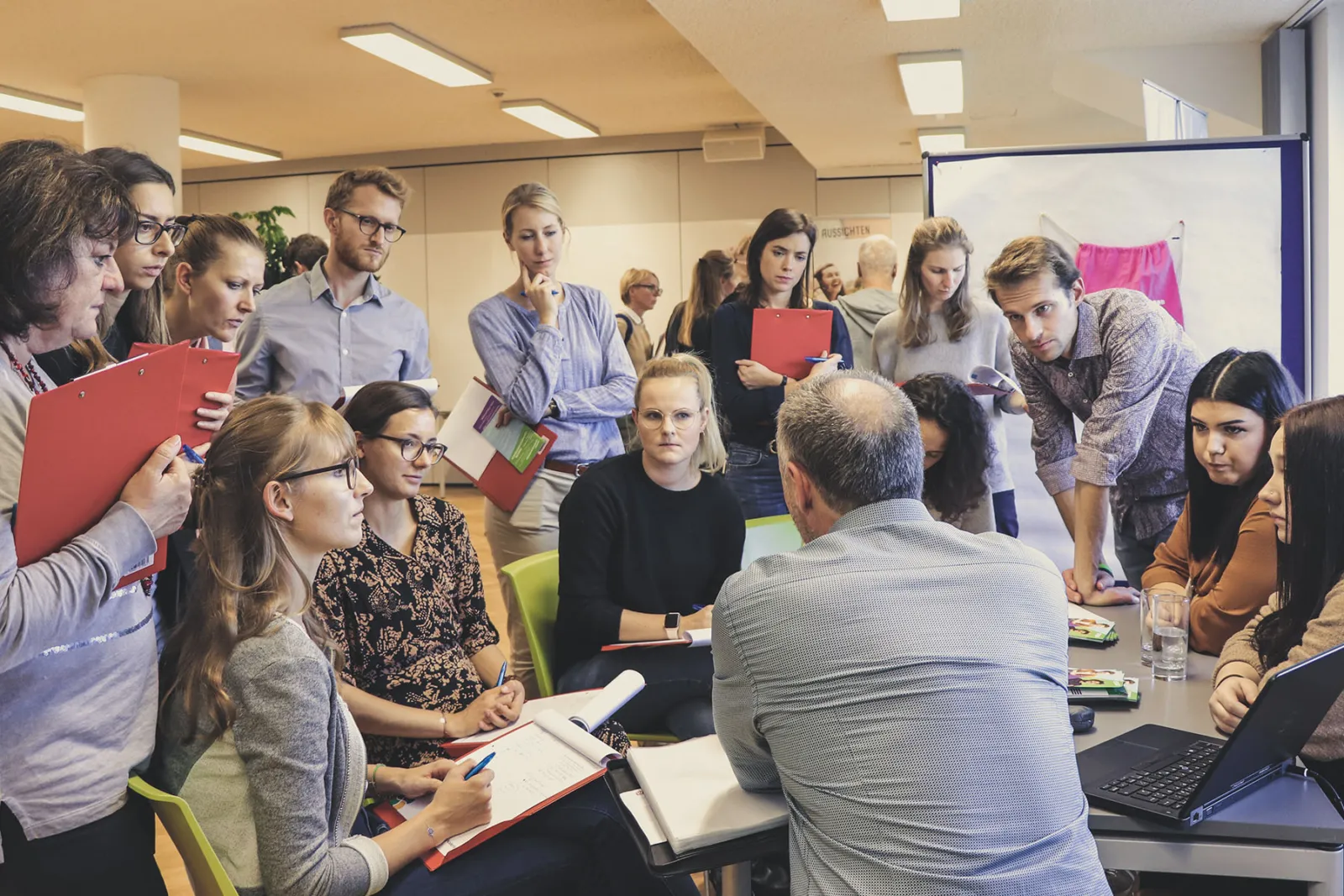 Arbeitsgruppe diskutiert in Konferenzraum um einen Tisch; Moderator erläutert, Notizblöcke, Laptop und Aktenordner liegen bereit.