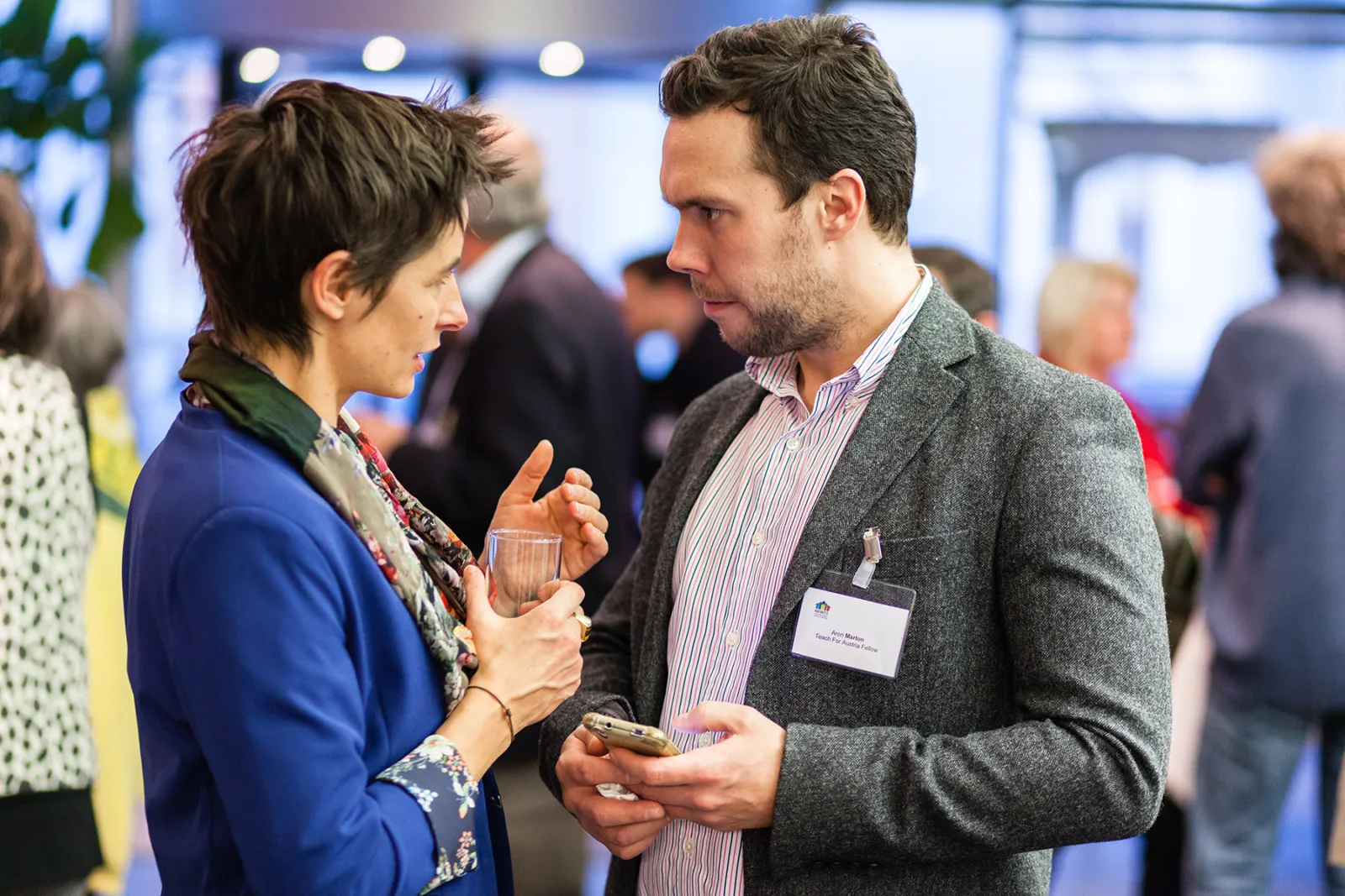 Zwei Fachleute im Gespräch auf einer Networking-Veranstaltung; eine Person hält ein Glas, die andere ein Smartphone.