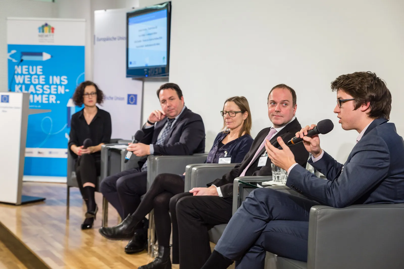 Fünf Panelteilnehmer diskutieren in einem Konferenzraum; rechts sitzender Panelteilnehmer hält Mikrofon, weitere Zuhörer lauschen.