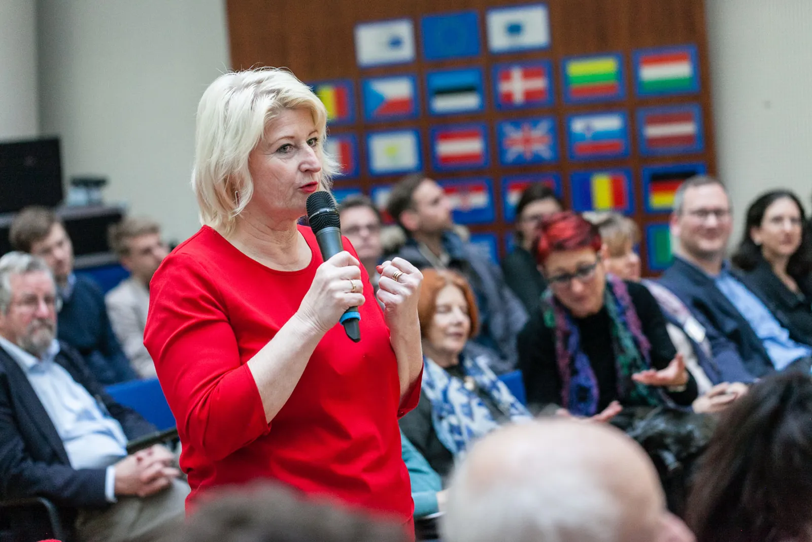 Person mit blonden Haaren in rotem Oberteil spricht ins Mikrofon vor Publikum bei einer Konferenz, Flaggenwand im Hintergrund.