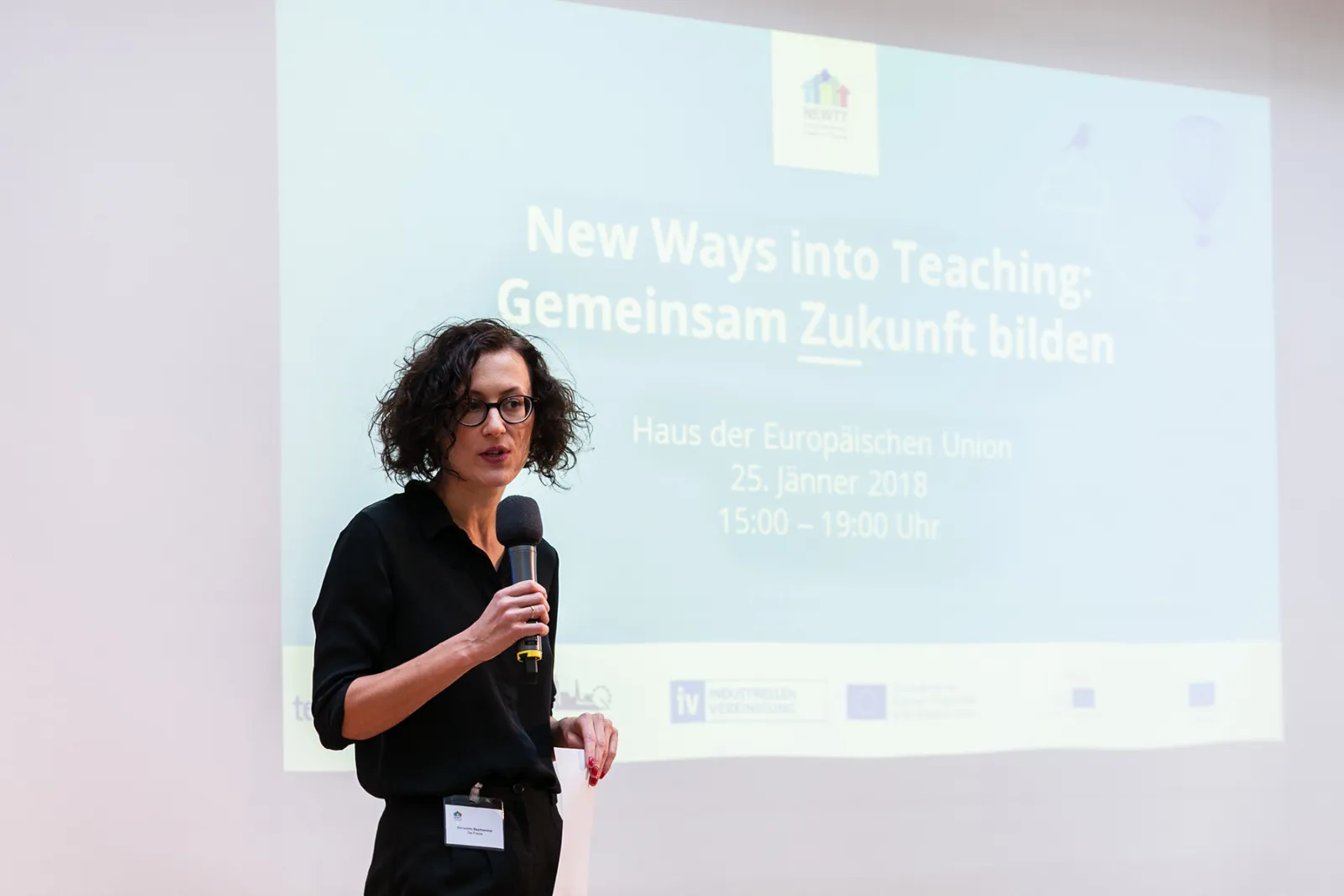 Frau mit dunklem Hemd und Brille hält Mikrofon vor projizierter Folie mit Veranstaltungsinfos, Haus der Europäischen Union.