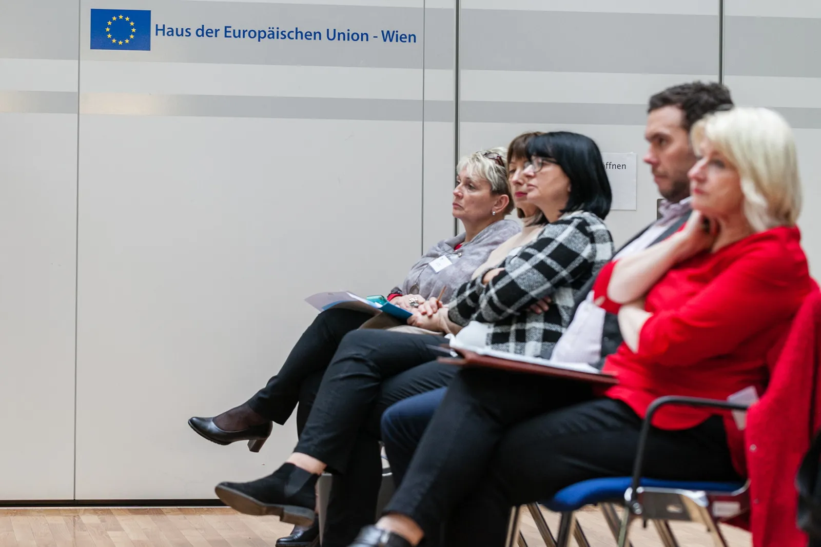 Gruppe von Erwachsenen sitzt aufmerksam in Sitzreihen, mit Notizblättern, im Raum des Hauses der Europäischen Union – Wien.
