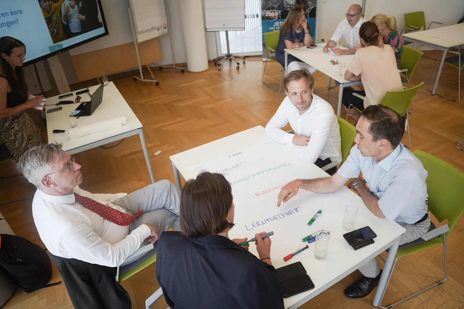 Gruppe von Geschäftsleuten sitzt an einem weißen Tisch, diskutiert und notiert mit Markern; Konferenzraum im Hintergrund.