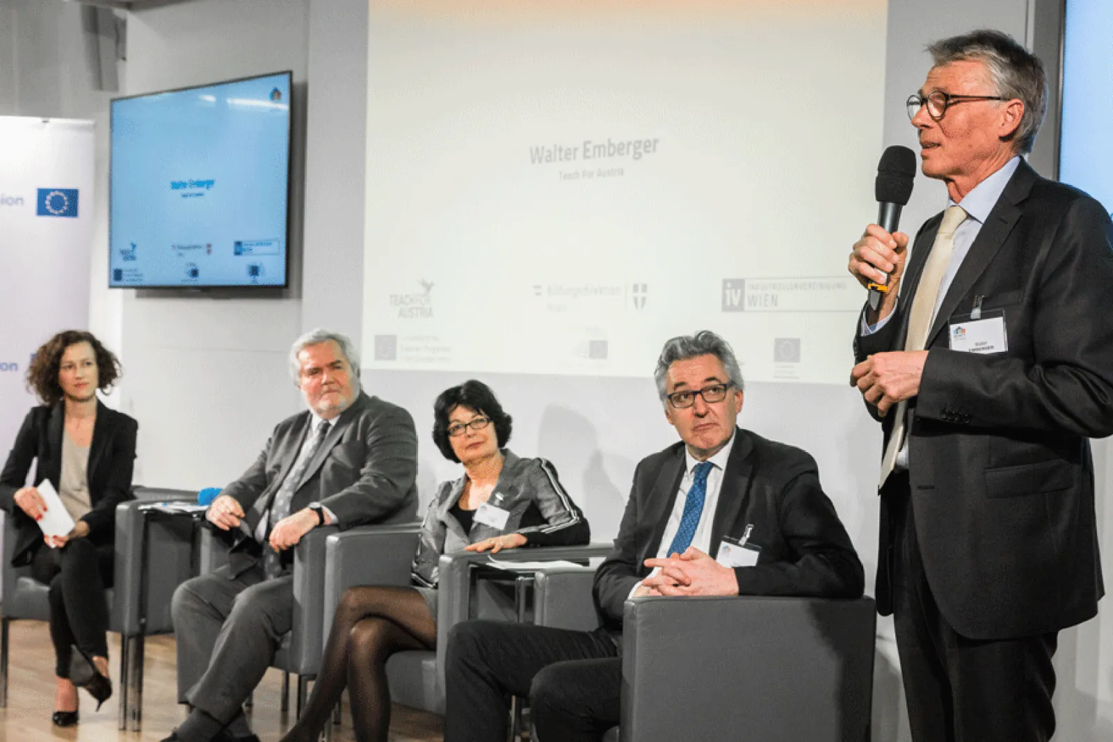 Sprechender Mann im Anzug mit Mikrofon bei Paneldiskussion; Panelteilnehmer sitzen im Konferenzraum.