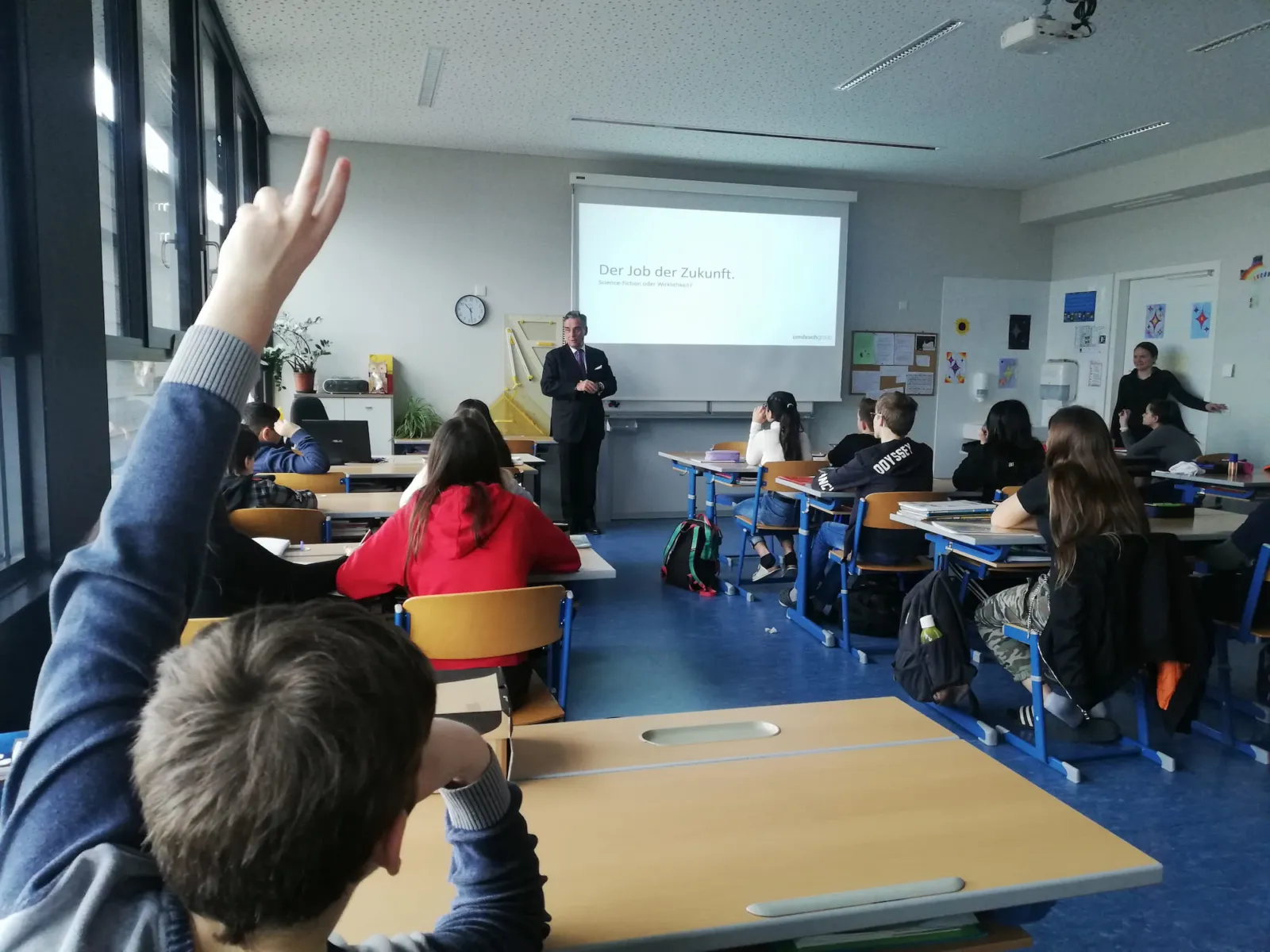 Dozent präsentiert vor Klasse; eine Person hebt die Hand; Projektion zeigt 'Der Job der Zukunft'.