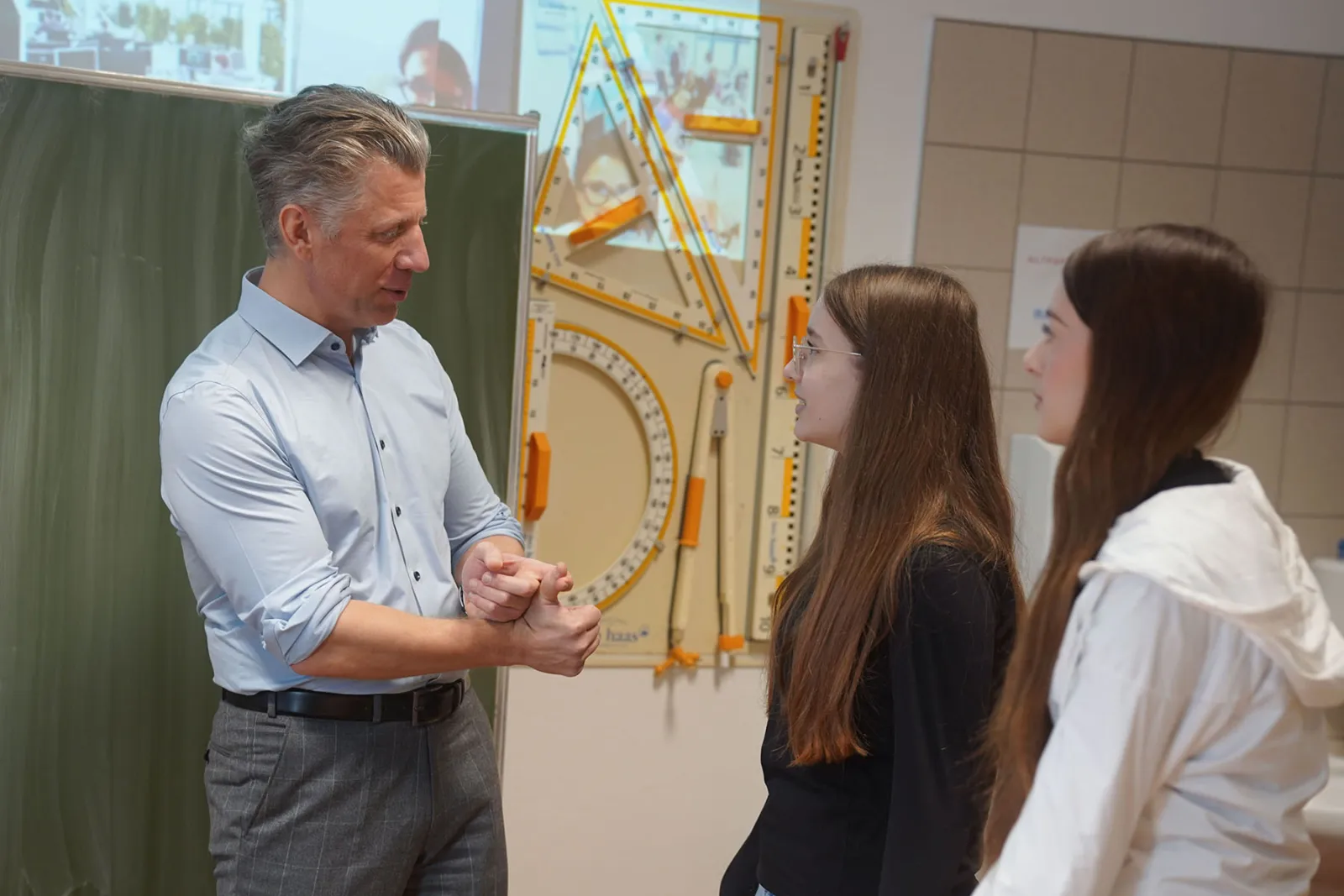 Lehrer spricht mit zwei Schülerinnen in Klassenzimmer; im Hintergrund große Geometrie- und Messinstrumente an der Wand.