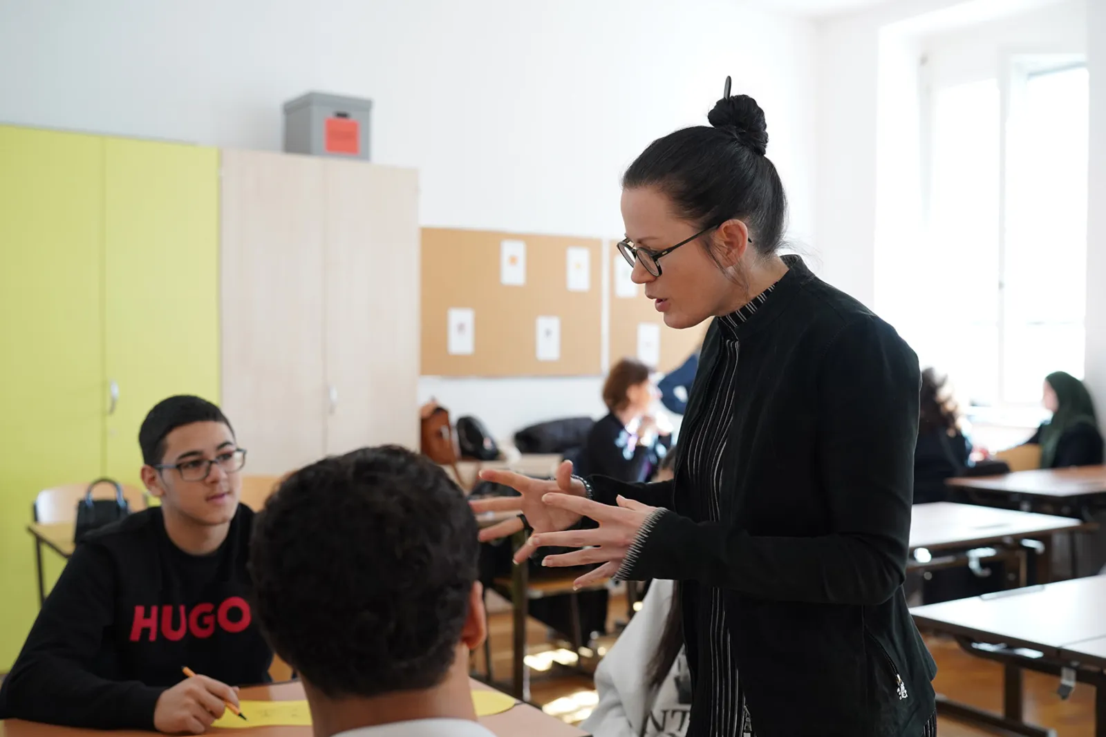 Lehrperson mit Brille gestikuliert und erklärt einer Lernperson an einem Tisch im Klassenzimmer; weitere Lernende arbeiten im Hintergrund.