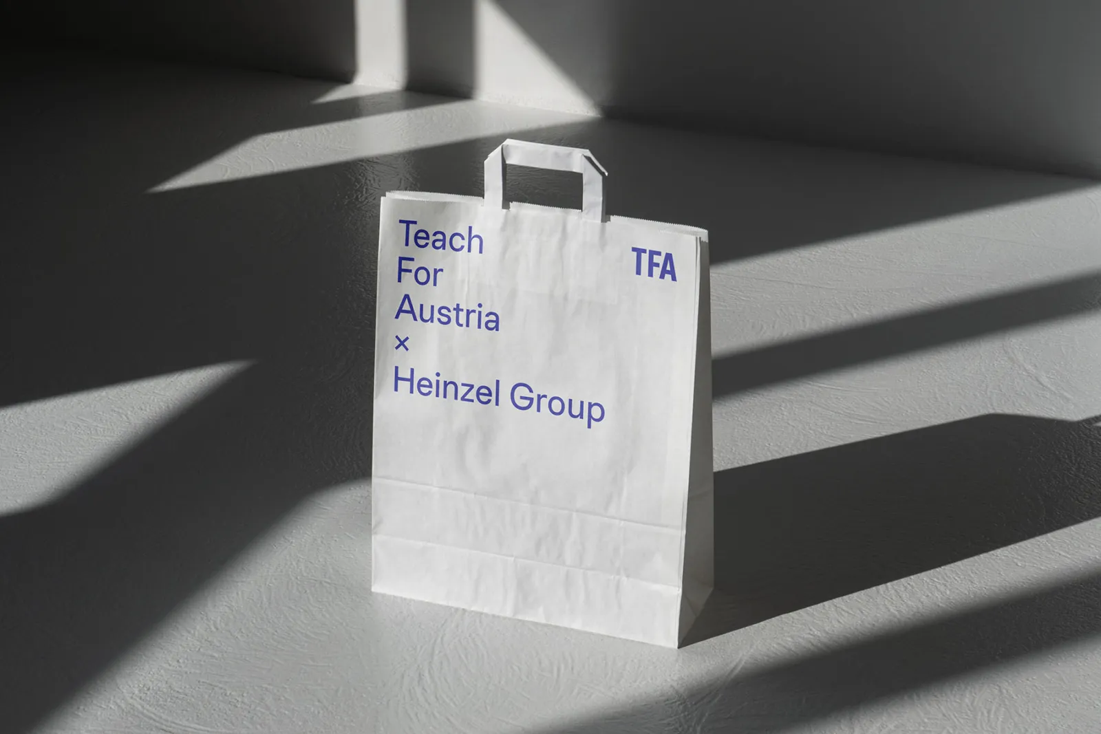 Weiße Einkaufstasche mit blauer Aufschrift 'Teach For Austria x Heinzel Group' steht auf grauem Boden; Licht- und Schattenmuster.