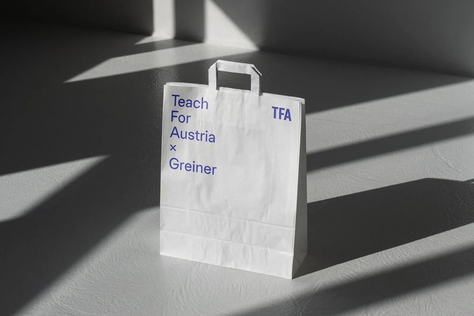 Weiße Papiertragetasche mit blauer Aufschrift 'Teach For Austria x Greiner' (TFA), steht auf grauem Boden.