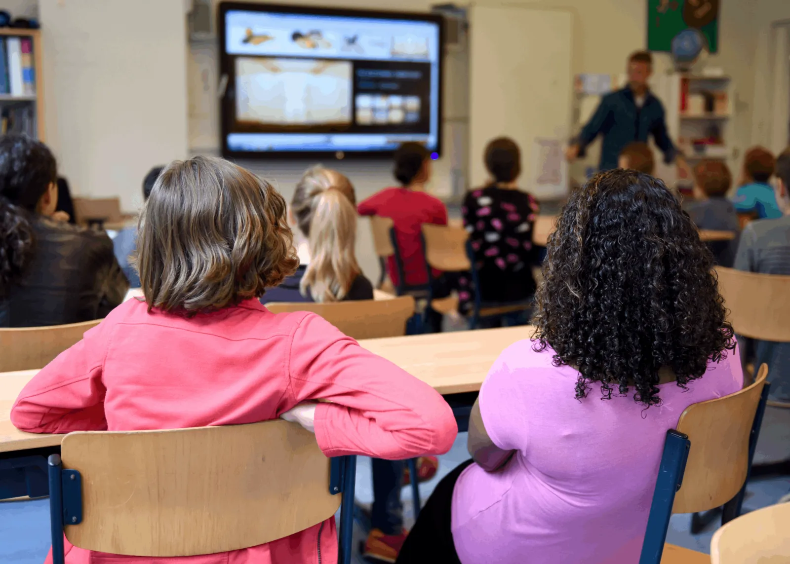 Zwei Lernende sitzen in einem Klassenraum, während eine Lehrkraft vorne eine Präsentation am Smartboard zeigt.