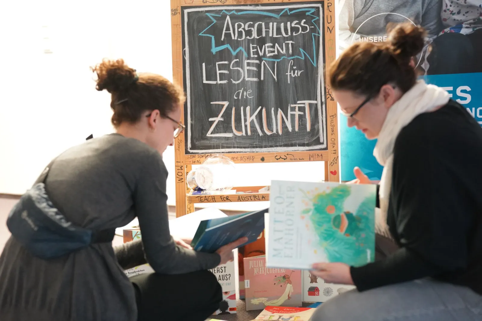 Zwei Personen lesen Bücher vor einer Tafel mit dem Text 'Abschluss-Event: Lesen für die Zukunft' bei einer Indoor-Veranstaltung.