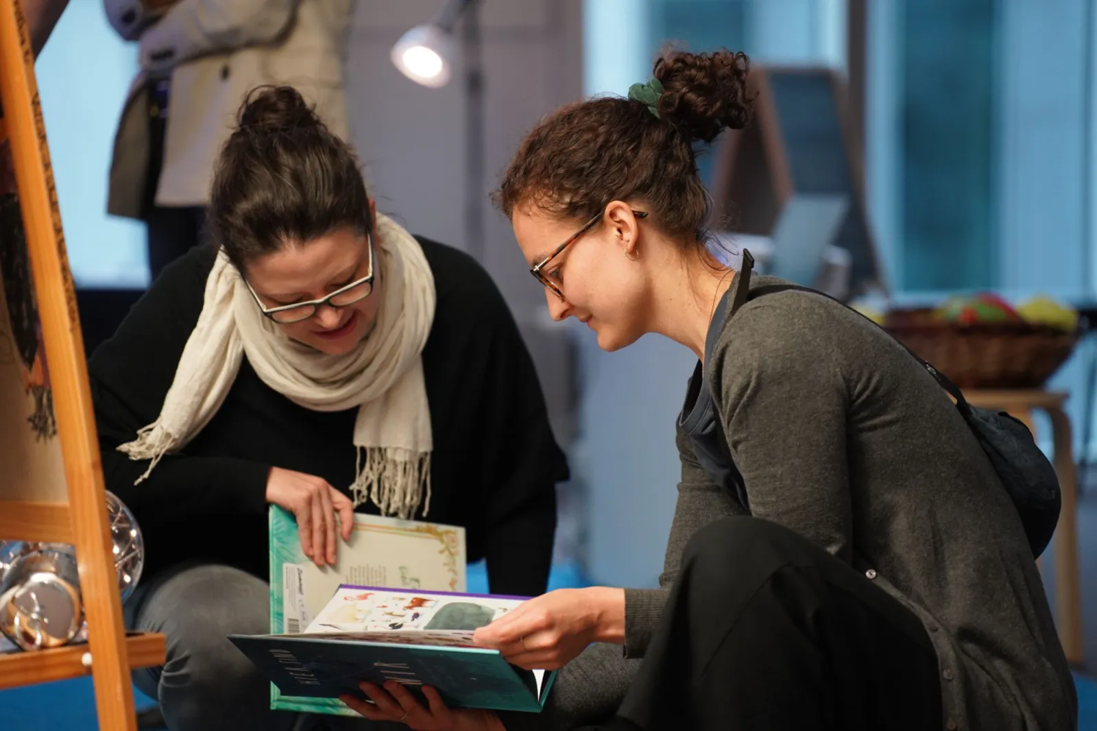 Zwei Personen sitzen auf dem Boden und betrachten gemeinsam ein Buch; im Hintergrund eine Staffelei.