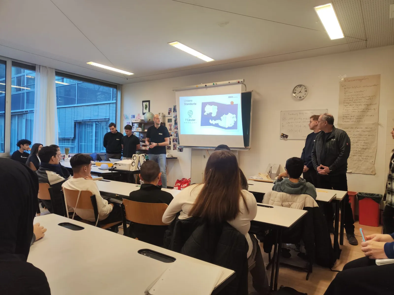 Klassenzimmer-Szene: Schülerinnen und Schüler hören Lehrkräften zu; Projektion einer Karte an der Wand.