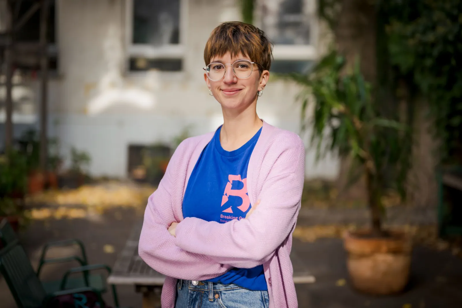 Person mit Brille, kurzem Haarschnitt, rosa Cardigan und blauem T-Shirt, verschränkte Arme, lächelnd im Gartenhof.
