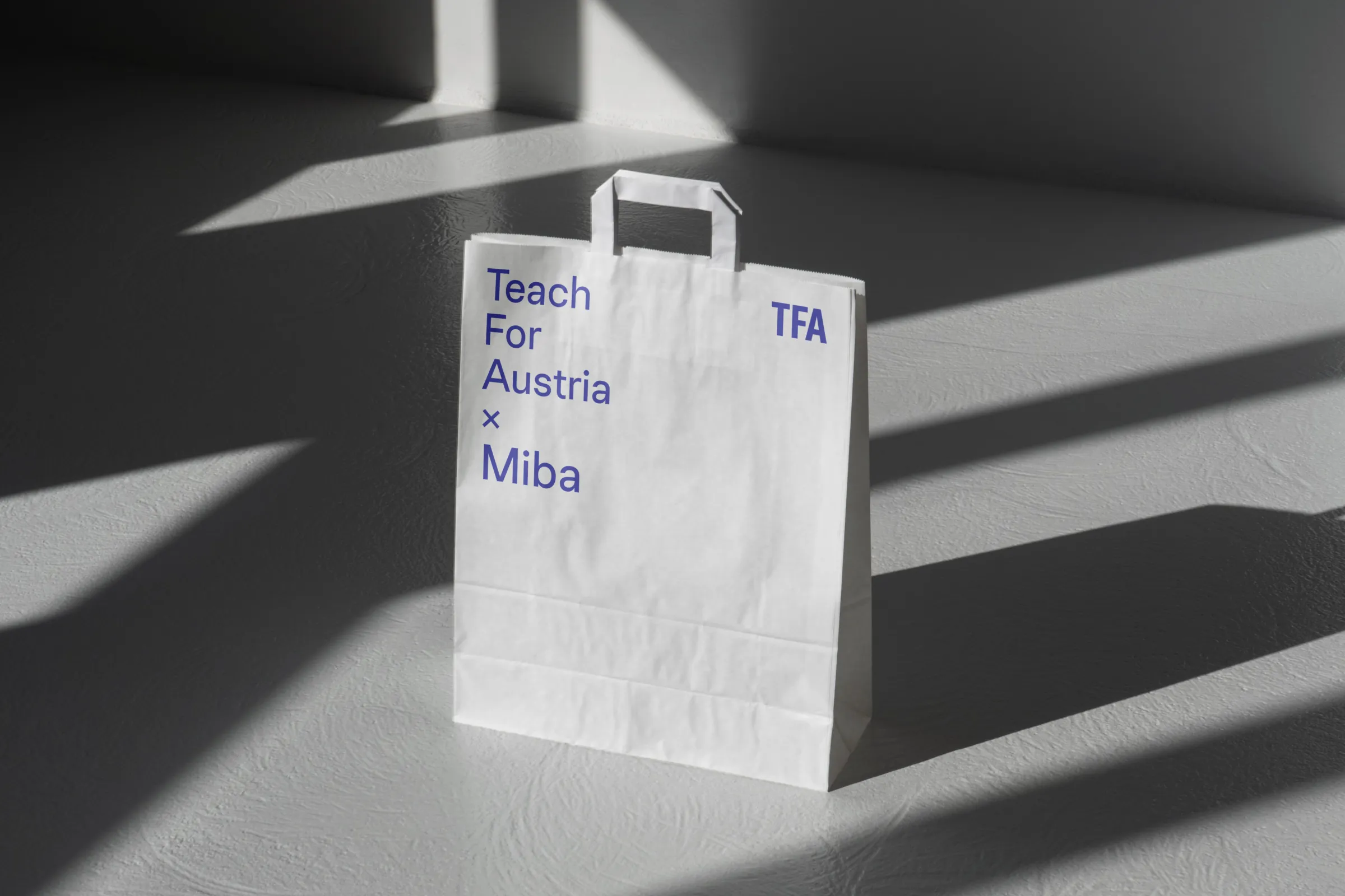 Weiße Papiertasche mit blauer Aufschrift 'Teach For Austria x Miba' steht auf grauem Boden; Lichtstreifen und Schatten im Raum.
