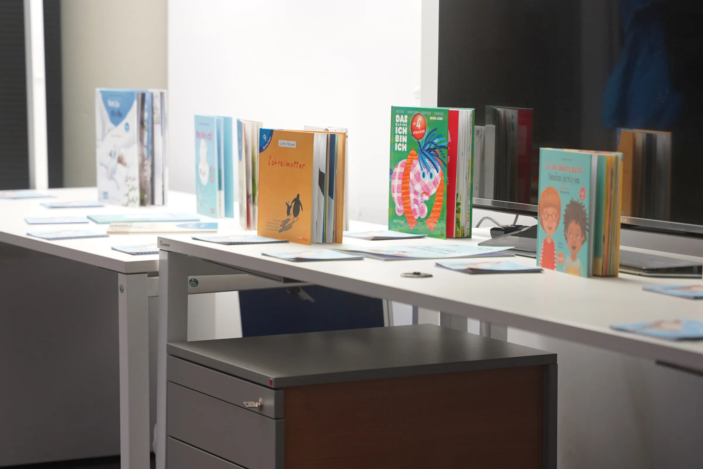 Bunte Kinderbücher stehen in einer Reihe auf einer weißen Tischplatte; im Hintergrund ein Bildschirm.