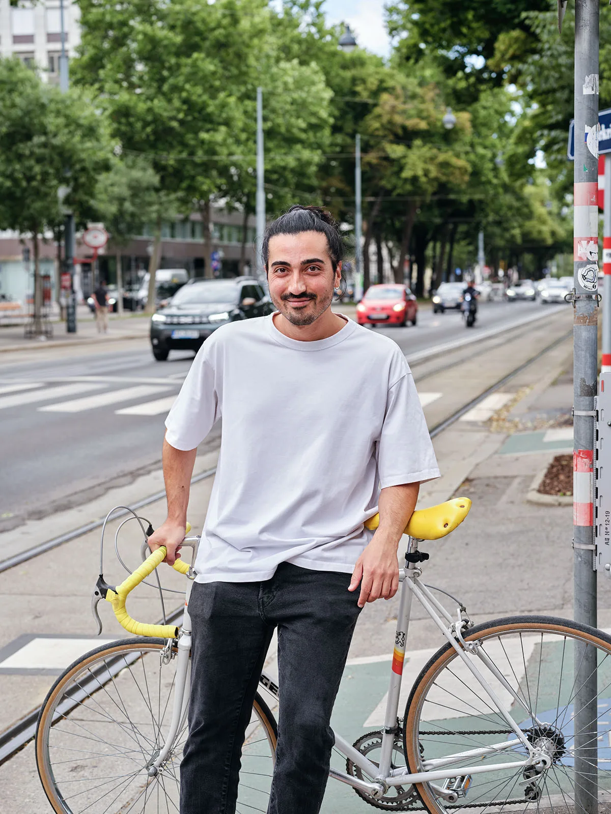 Person steht lächelnd neben hellgrauem Rennrad mit gelber Sattel und gelbem Lenkerband auf urbaner Straßenecke mit Straßenbahnschienen.