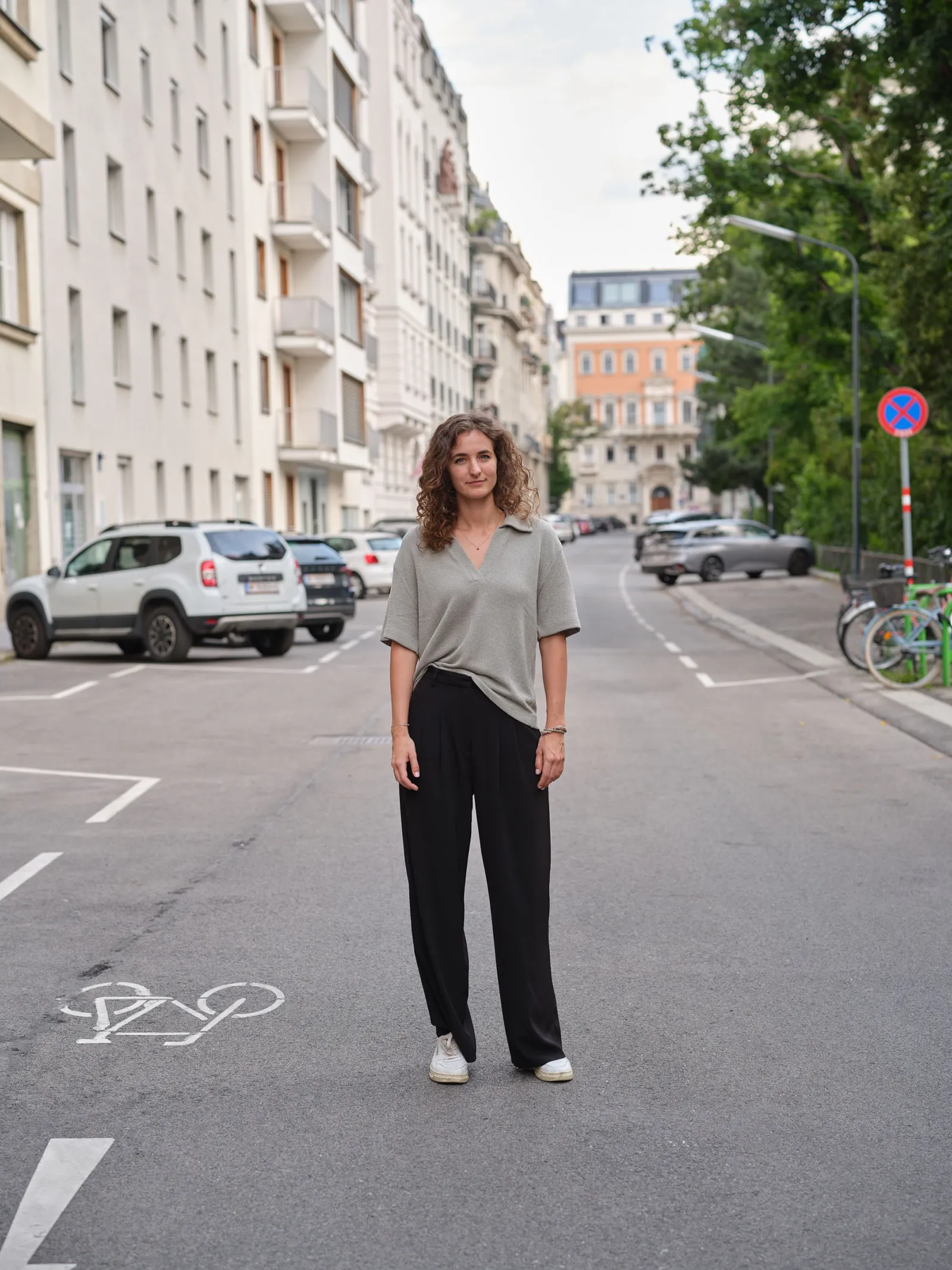Person mit lockigen Haaren steht mitten auf einer Stadtstraße, graues Oberteil, schwarze weite Hose, weiße Sneaker; parkende Autos sichtbar.