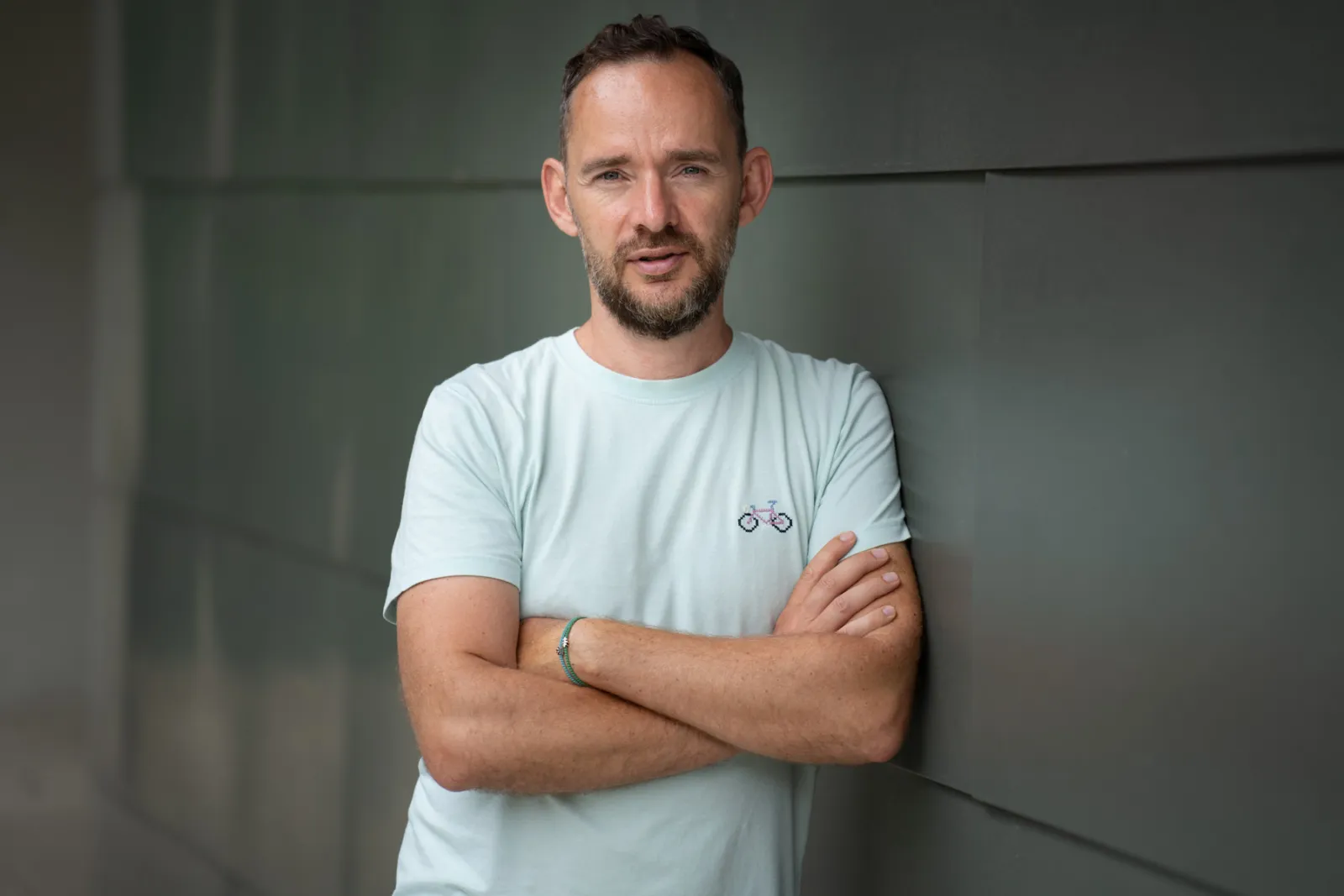 Mann mit kurzem Bart lehnt an einer grauen Wand, Arme verschränkt, mintfarbenes T-Shirt.