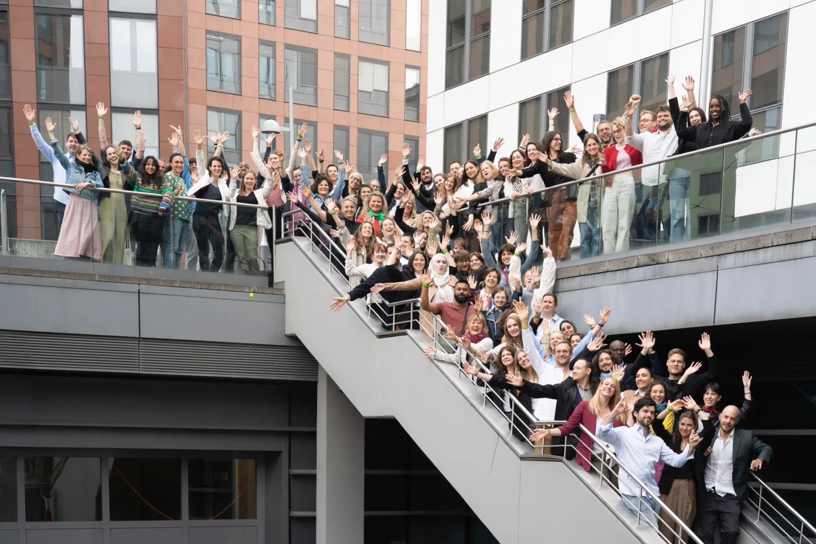 Große Gruppe von Menschen winkt von Treppe und Balkon eines modernen Bürogebäudes in die Kamera.