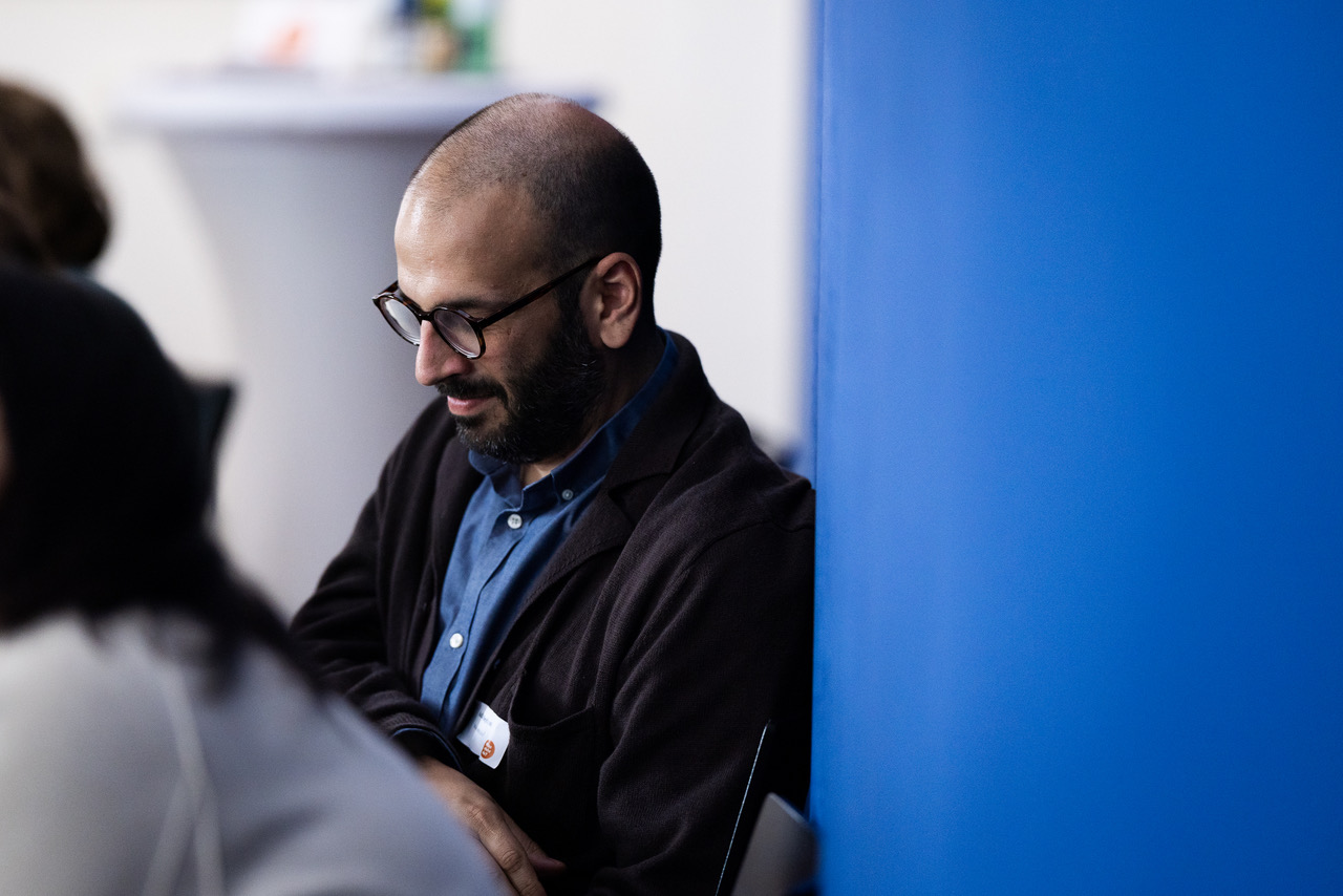 Person mit Brille und Bart sitzt an blauer Wand, schaut nach unten; trägt blaues Hemd und dunkle Jacke.