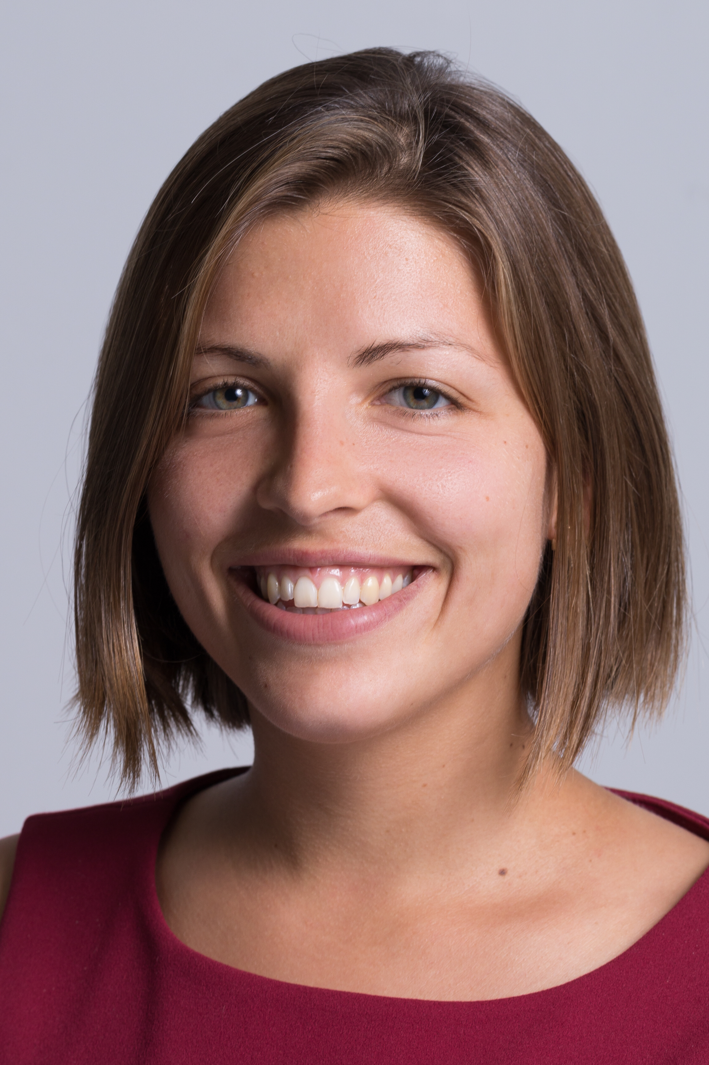 Porträt einer Person mit braunen kurzen Haaren, lächelnd, burgunderrotem Oberteil vor neutralem Hintergrund.