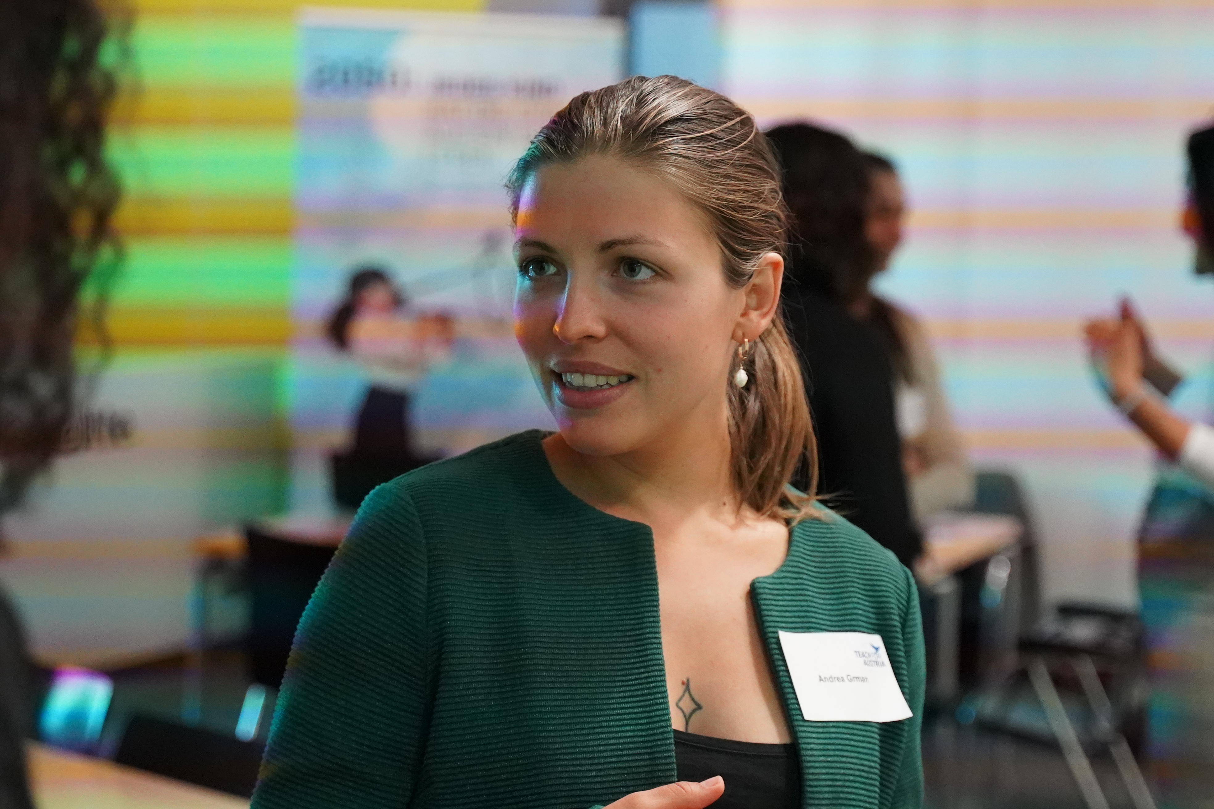 Person in grünem Blazer spricht auf einer Veranstaltung; Namensschild sichtbar, Hintergrund mit bunten Streifen und weiteren Teilnehmenden.