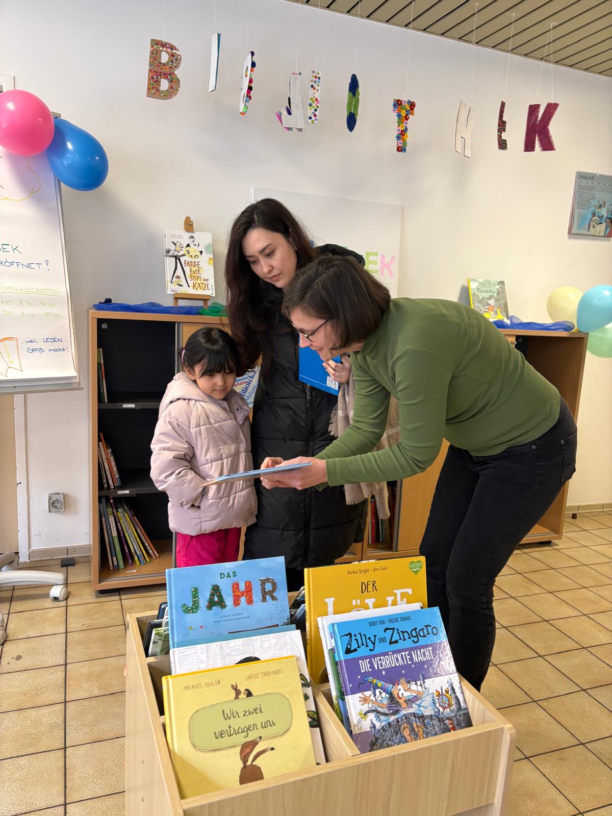 Person im grünen Pullover zeigt einem Kind im Lesesaal ein Blatt; daneben stehen zwei Erwachsene, vorne Bücherstapel.
